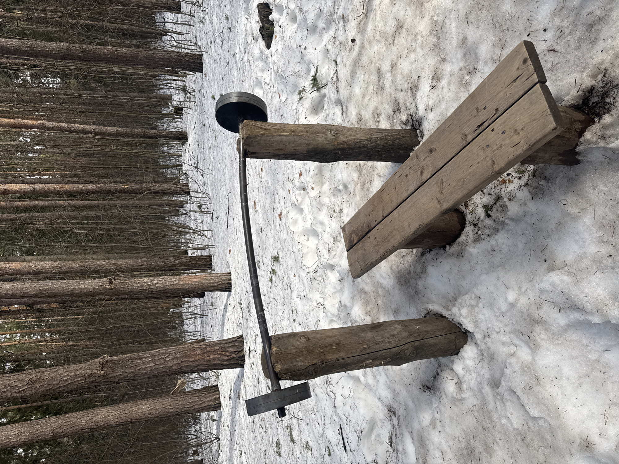The scene is an outdoor exercise area, likely located within a park or wooded area in Votkinsk, Russia, given the prompt's information. The ground is partially covered with melting snow. The exercise equipment consists of a wooden bench supported by logs and a weightlifting apparatus made from logs, a metal bar, and weight plates. There are no people visible in the image. The background shows a dense forest of bare trees.