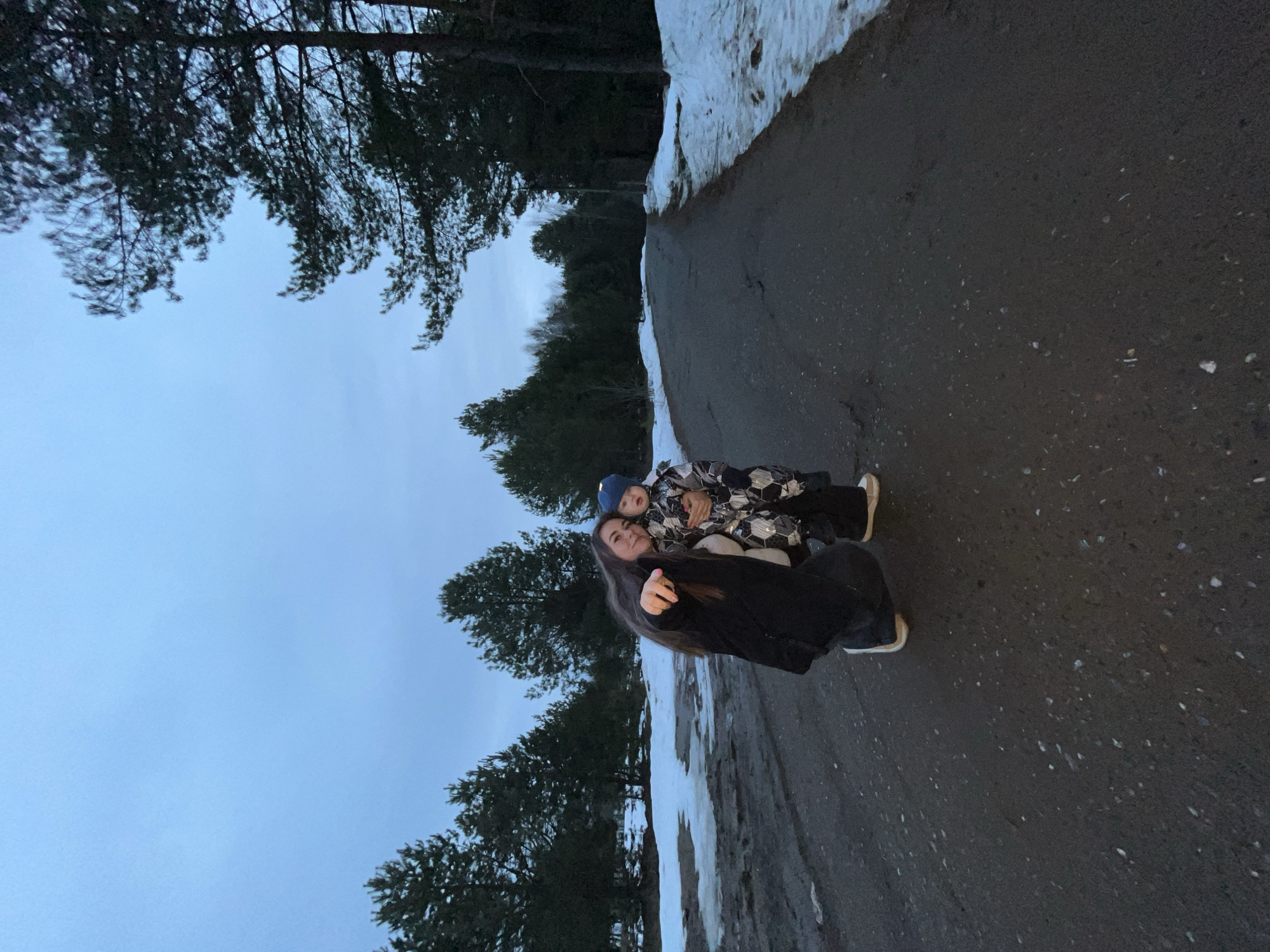 The image depicts a woman and a child standing on a road. The road is lined with snow on one side, and trees are visible in the background and to the left. The sky is overcast. The woman is wearing dark clothing and holding the child. The child is wearing a patterned jacket and a blue hat. The location appears to be a road in a wooded area, possibly in the city of Chaikovsky, Russia.