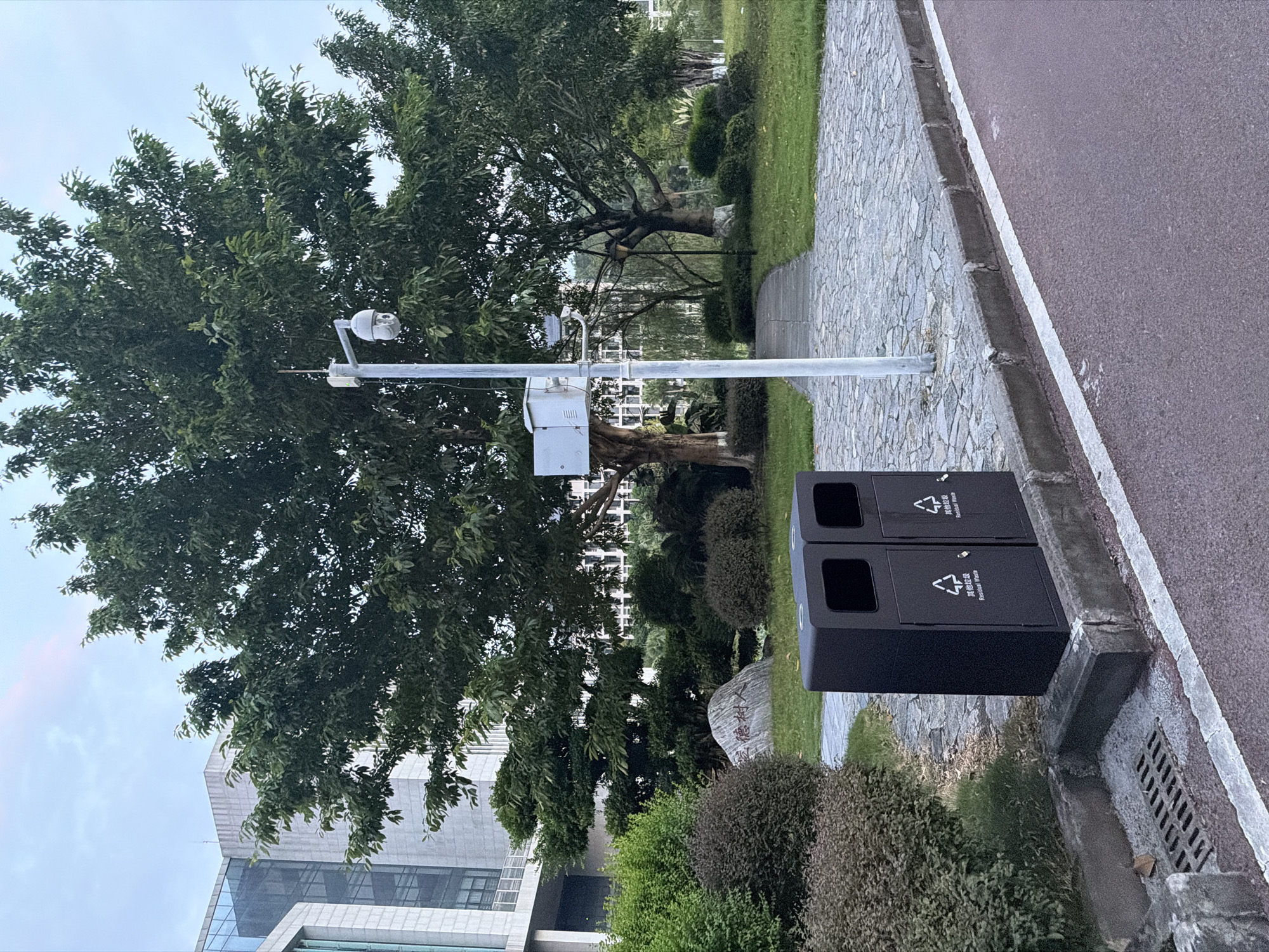 The image depicts an outdoor scene in Jiangjin, China, likely a public space such as a park or campus, characterized by a mix of natural and man-made elements.
In the foreground, two black trash receptacles stand side-by-side on a gray concrete curb, adjacent to a textured red surface, possibly a running or biking path. Each receptacle has two openings at the top and a compartment at the bottom. Chinese characters and the English phrase "Residual Waste" are visible on the front of the bins. A metal grate for drainage is located to the left of the trash cans, embedded in the concrete.
Behind the trash cans, a stone pathway made of irregular gray flagstones winds through a landscaped area. This area features various types of green foliage, including manicured bushes and trees. A large, light-colored rock with Chinese characters inscribed on it sits within the greenery.
A prominent feature in the midground is a tall, slender, light-colored pole supporting a security camera and what appears to be an electrical box. The camera is a white, dome-shaped unit mounted on an adjustable arm. The pole is situated near a mature tree with a thick trunk and lush green leaves.
In the background, a modern building with large glass windows and a light-colored facade is visible, suggesting an institutional or commercial setting. The sky above is overcast with a pale pinkish hue, indicating it may be dusk or dawn. The overall atmosphere is serene and orderly.