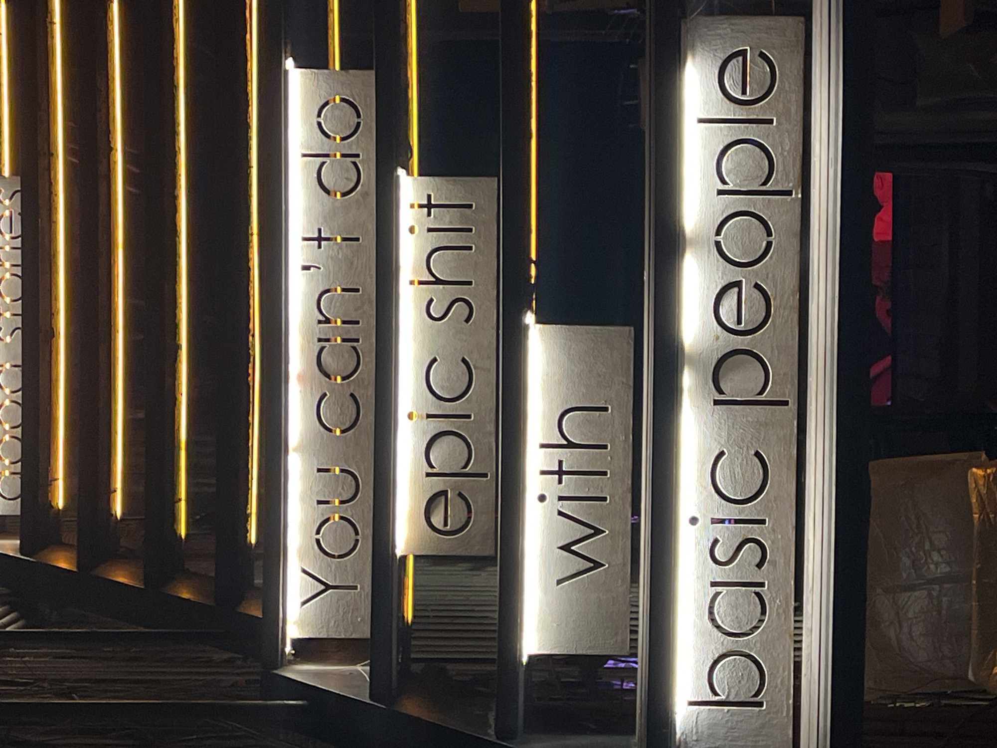 This media file depicts a nighttime scene in Baan Bo Phut, Thailand, featuring a decorative display of illuminated vertical signs. The signs are made of a material that reflects light, creating a glowing effect. Each sign contains text cut out in a stylized font, allowing the internal lighting to shine through.
The visible text on the signs reads as follows, from left to right: "You can't do", "epic shit", "with", and "basic people". The arrangement of these phrases suggests a motivational or humorous message.
The overall setting appears to be an outdoor area, possibly a street, walkway, or entertainment venue. The background is dark, consistent with nighttime, and is punctuated by yellow vertical lights that create a striking contrast with the illuminated signs. Some areas in the background are out of focus, but hints of structures and possibly wrapped objects can be seen. The lighting is dramatic and creates a modern, somewhat edgy ambiance. There are no people visible in the frame. The weather is not discernible from the image.
