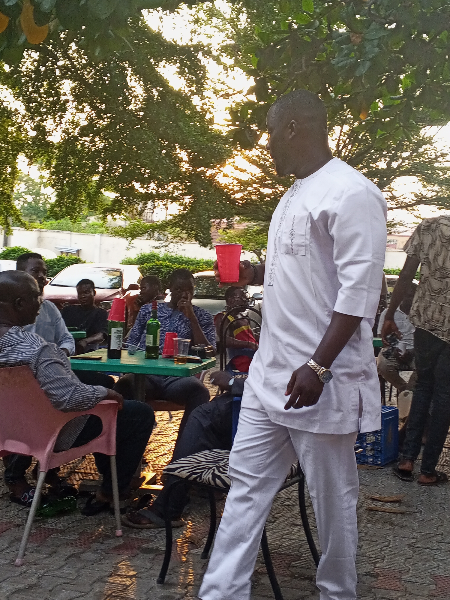 In Lokoja, Nigeria, the image captures an outdoor social gathering at dusk. A group of approximately seven people is seated around two adjacent green tables. One table is laden with a wine bottle and red plastic cups. A man dressed in a white, formal-style shirt and pants stands prominently in the foreground, holding a red cup. Several individuals are seated at the tables, engaged in what appears to be conversation. The setting is an outdoor space, presumably a patio or gathering area, with trees in the background. A vehicle is parked nearby. An empty blue plastic crate is placed on the ground near the edge of the gathering.