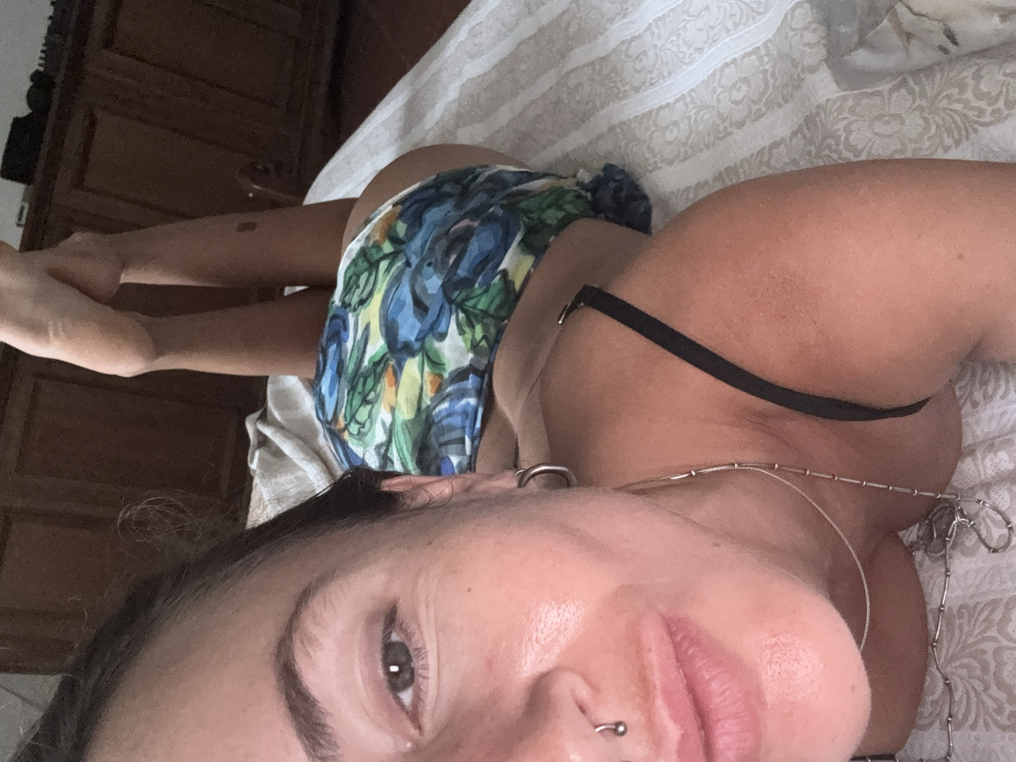 A selfie is taken from a close-up, high-angle perspective of a person lying on their back on a bed. The subject's face is in the foreground, with their left eye, nose, and mouth visible. They have dark hair, a septum piercing, and a hoop earring. The subject is wearing a colorful, floral-print garment with a black strap visible on their shoulder. Their legs are extended upwards in the background. The environment appears to be a bedroom, with a wooden dresser visible behind the subject and a patterned bedspread. The lighting is soft, suggesting an indoor setting.