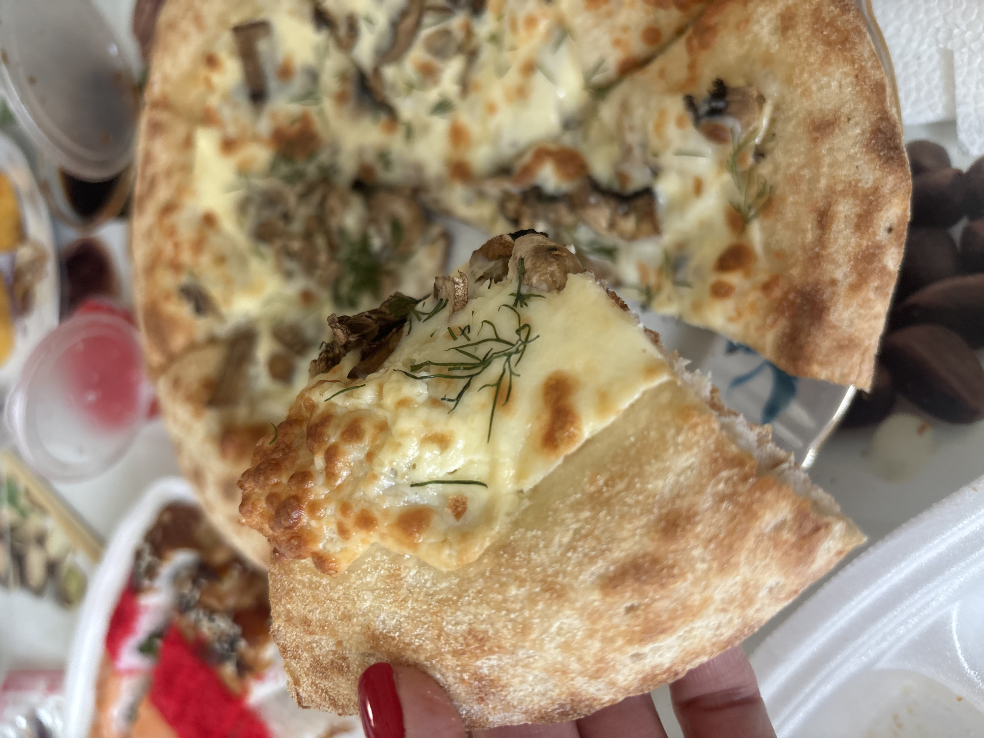 A slice of mushroom pizza is being held up by a hand with red nail polish, with the rest of the pizza visible in the background. The pizza has a light-colored crust, white cheese, mushrooms, and dill. Other food containers are visible in the periphery, including a clear container with a red liquid. The location is Chaikovsky, Russia.