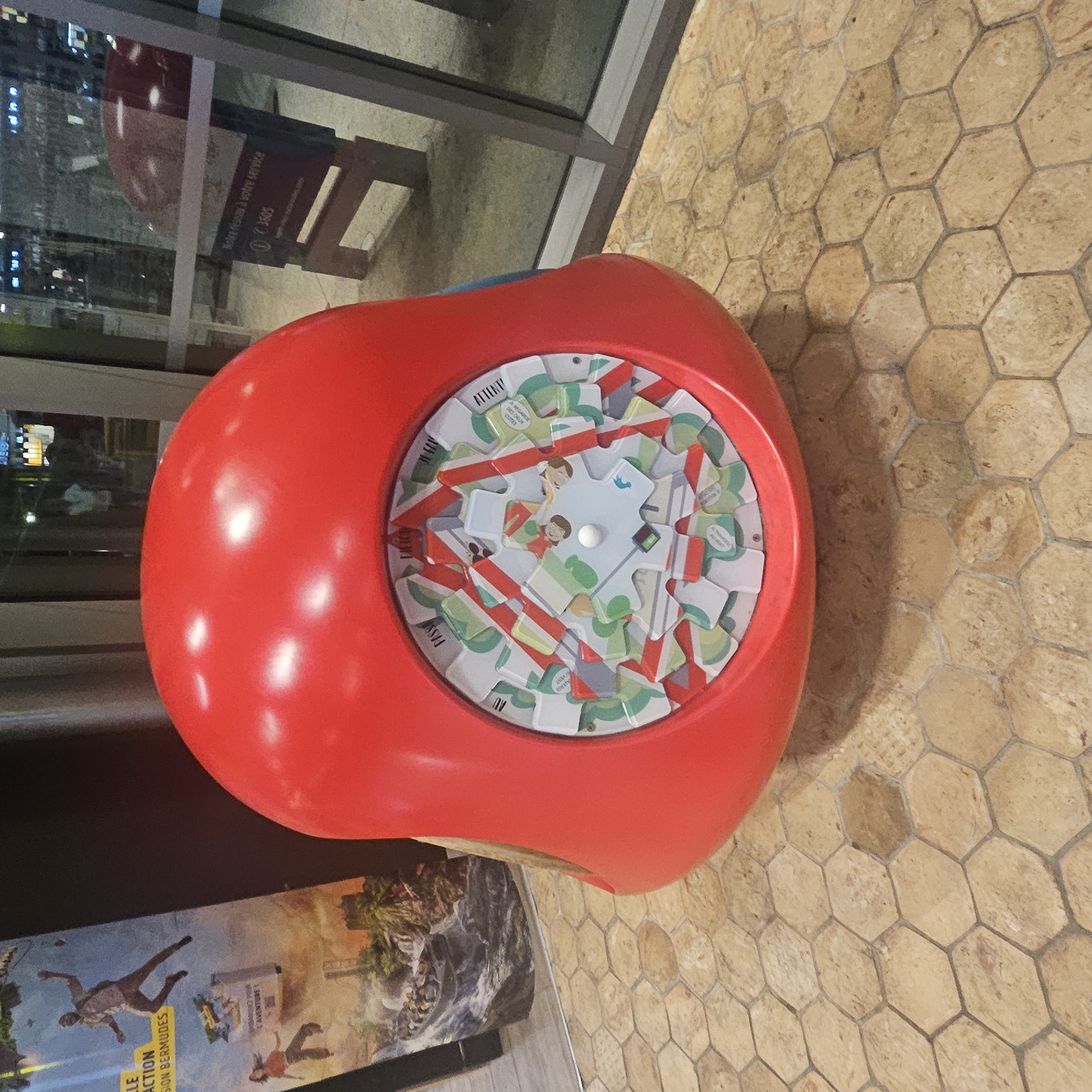 The image shows a red, mushroom-shaped children's play structure with a circular interactive element on its front. This interactive element features a colorful, rotating mechanism with various illustrated panels and gears. The structure is situated on a hexagonal tiled floor with a tan and brown pattern. In the background, there is a large window with reflections of the interior and exterior, including a sign that reads "Notre réseau à votre service 0 3605." To the left of the structure, a large poster is visible, depicting scenes of adventure with people in a boat on a waterfall and a person jumping, with text in French that appears to be related to travel or an attraction. The scene appears to be indoors, possibly in a public space like a train station or visitor center, given the signage and the reflective surfaces. The provided context indicates the location is Lançon-Provence, France.