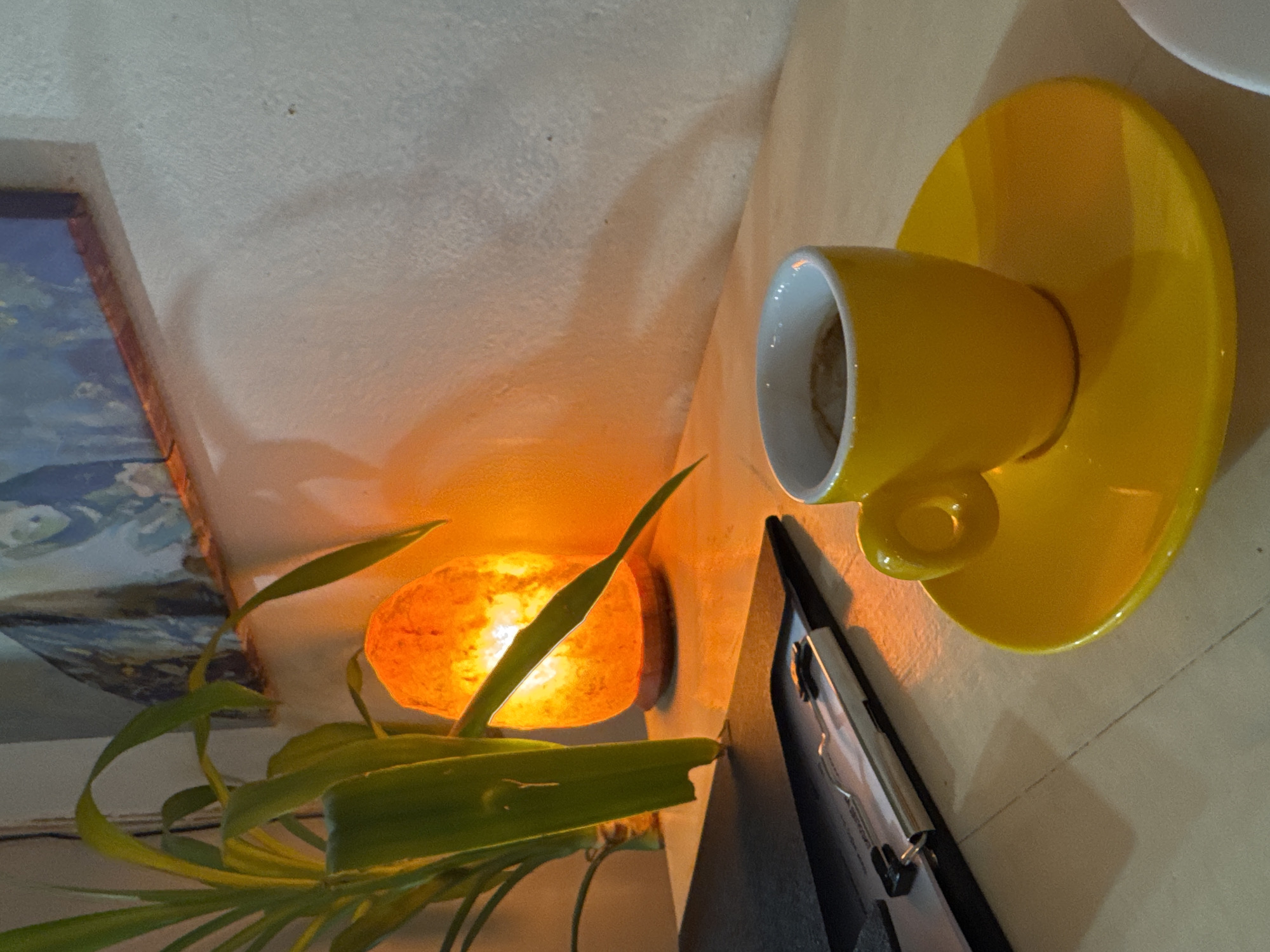 A vertical photograph captures a cozy indoor still life, rich in warm artificial light, set in an unspecified location within Sofia, Bulgaria. The camera is tilted, presenting the scene as if viewed from above and to the side, with objects arranged on a light-colored surface.
The focal point on the right is a vibrant yellow ceramic coffee cup resting on its matching saucer. The cup's interior is white, showing a slight residue, suggesting it has been recently used. Its cheerful color provides a contrast to the muted background.
Towards the center, a luminous Himalayan salt lamp emits a soft, amber glow, casting gentle, elongated shadows on the textured, off-white wall behind. This warm light source creates an inviting and tranquil atmosphere. Partially obscuring the lamp and extending from the lower left is a green potted plant with long, flowing leaves, adding a natural touch to the decor.
On the far left, the corner of a framed artwork or painting is visible, showcasing blue and white hues, possibly depicting a landscape or a cloudy sky. In the lower right corner, a dark-colored folder or clipboard with metallic clips is partly seen.
The scene conveys a sense of quietude and warmth, likely taking place in the evening or at night given the prominence of artificial light. There are no people visible, and no discernible text appears on any of the objects.