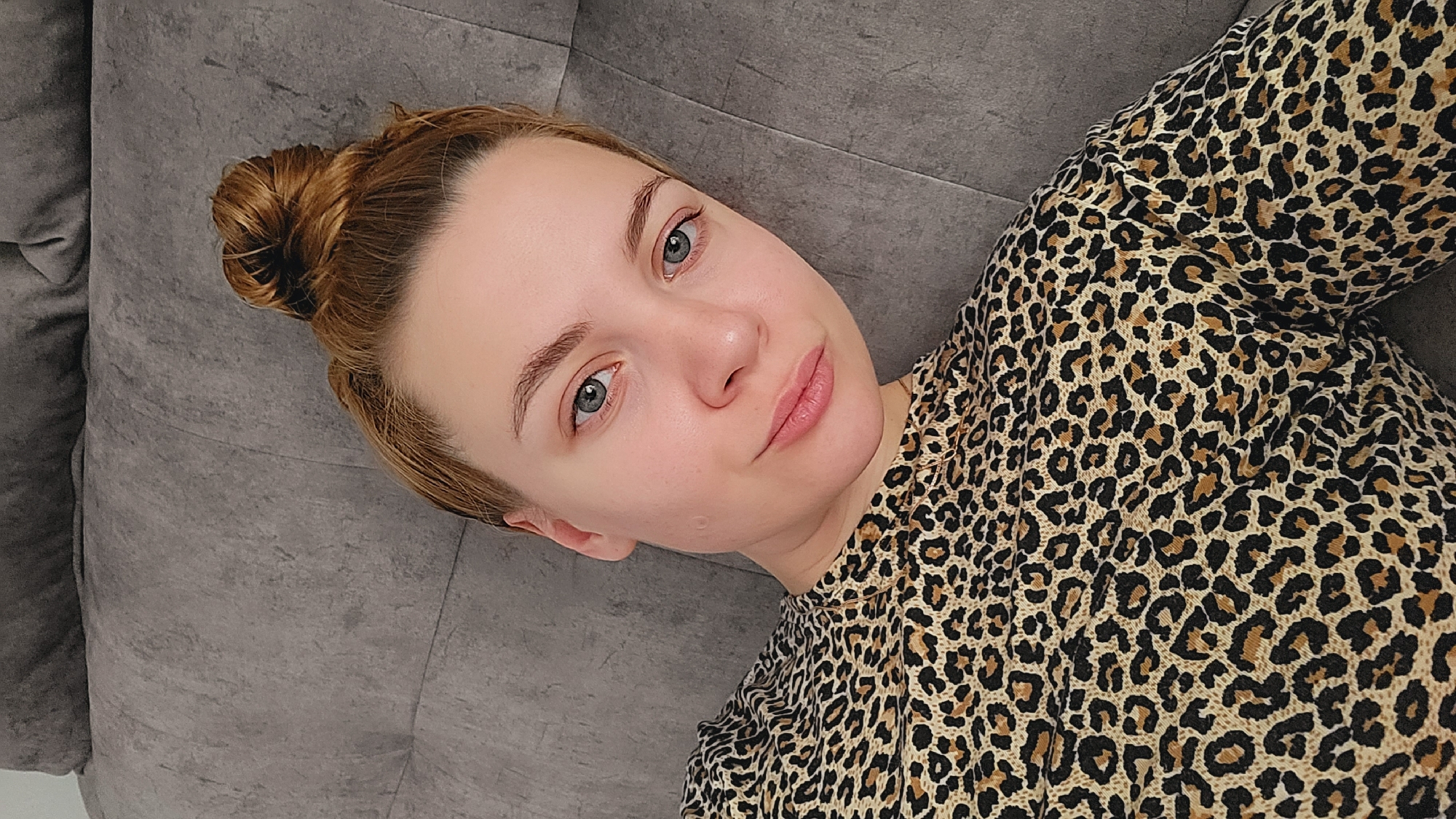 A woman with fair skin, light brown hair in a bun, and light blue eyes is lying on a gray upholstered surface, possibly a couch or bed. She is wearing a top with a brown and black leopard print pattern. The background is nondescript, suggesting an indoor location. It is not possible to identify the location as Chaikovsky, Russia, based on the image alone.
