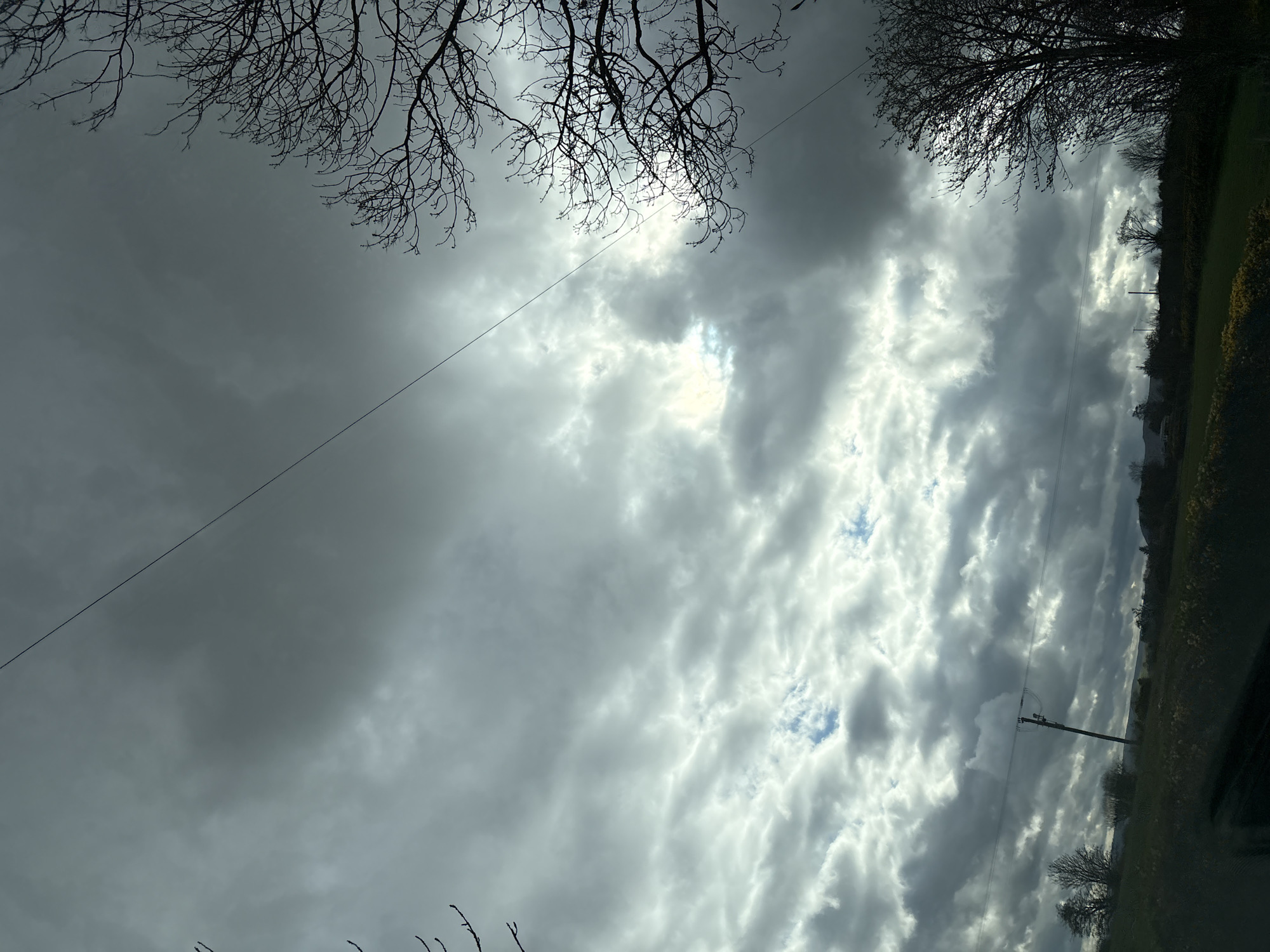 The image captures a cloudy sky with bare tree branches in the upper corners. A wire stretches diagonally across the left side of the frame. In the distance, a green field and a utility pole are visible on the right. The photo appears to have been taken in or near Stiperstones, United Kingdom, given the landscape and cloud formations.