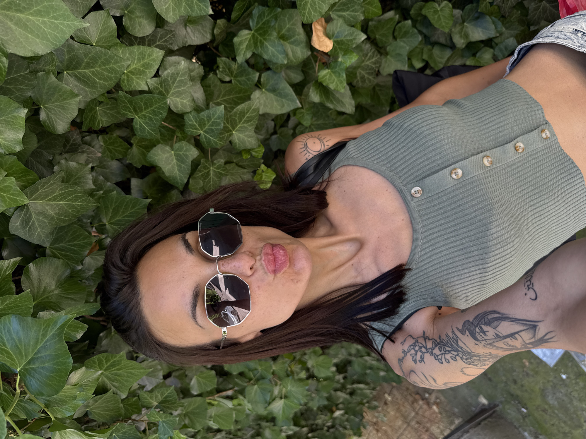 A woman lies among ivy plants. She has dark brown hair, wears sunglasses and a green button-down top. She has tattoos on her shoulder and arm. The photograph was possibly taken in a garden or park. The exact location in Pleven, Bulgaria cannot be determined from the image.