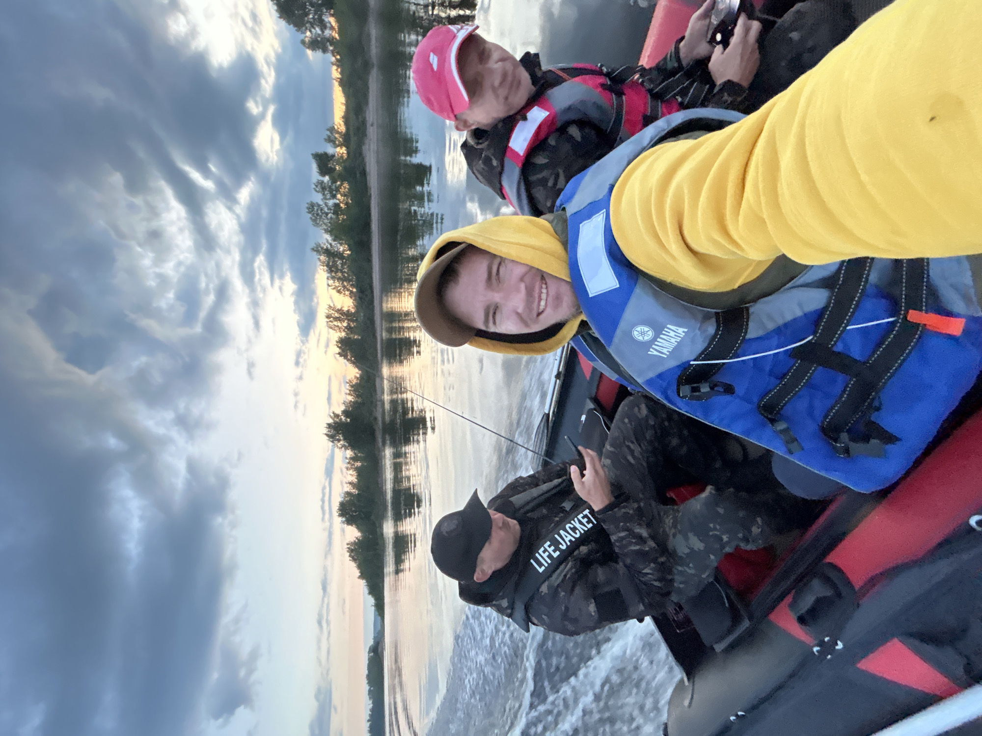 Three individuals are in an inflatable boat on a wide river at dusk. The sky is cloudy with hues of orange and pink near the horizon. Trees line the distant shore. The person in the foreground, wearing a yellow hoodie and a blue Yamaha life jacket, is taking a selfie. To their left, a person in a black cap and a camouflage jacket with "LIFE JACKET" printed on it is looking at their phone. To the right, a person in a red cap and a life jacket is visible. The boat is moving, creating a wake.