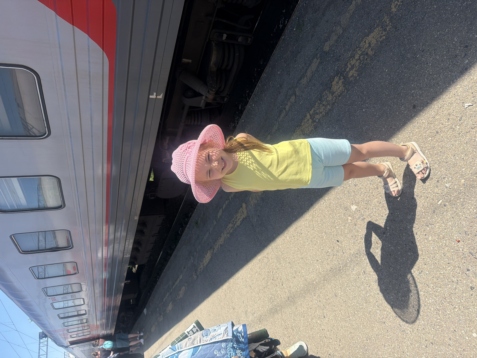 A young girl wearing a pink straw hat, a yellow t-shirt, and light blue shorts stands on a train platform in Kanash, Russia. She is smiling and looking at the camera. To her left, a train is parked on the tracks. The train is silver with red accents. The platform is made of asphalt and has yellow lines painted on it. The sky is clear and blue. The girl's shadow is cast on the ground to her left, shaped like a person with a wide-brimmed hat. There are other people and luggage visible in the background to the left of the frame.