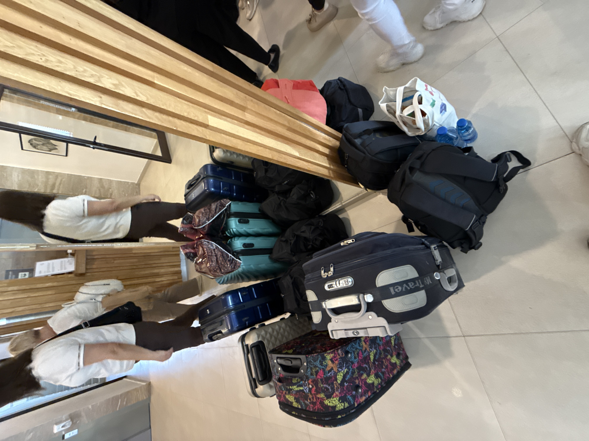 The image shows a collection of luggage placed near a slatted wooden structure in an indoor space, likely a waiting area or lobby. The luggage includes suitcases, backpacks, and bags of various colors and designs. There are also people in the image, some visible in the foreground near the luggage and others reflected in a nearby mirror. The flooring appears to be light-colored tile. The scene does not obviously indicate the location as Veliko Tarnovo, Bulgaria. Additional details such as specific architectural features, signage, or other contextual clues would be needed to confirm the location.