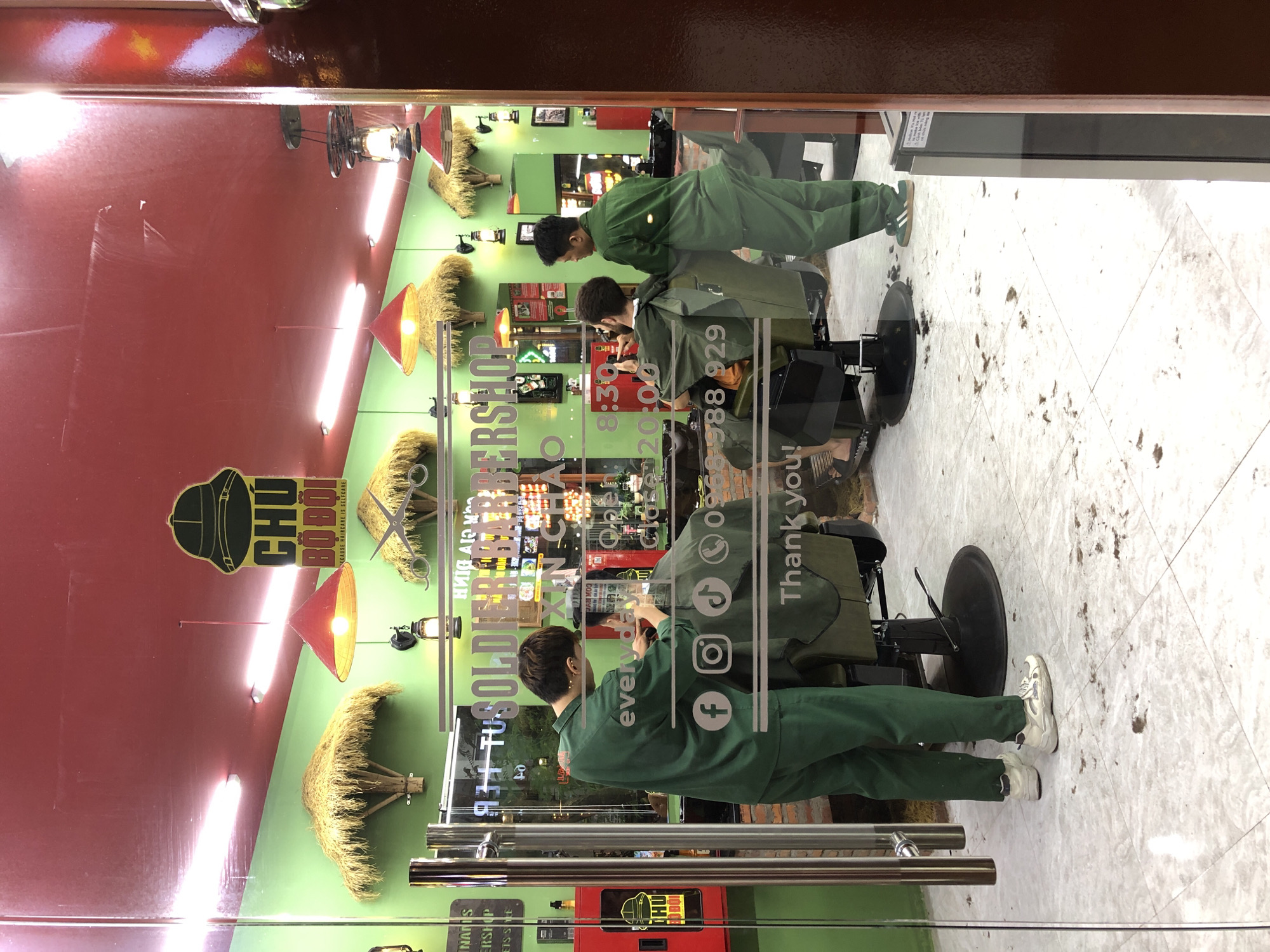 This is a full, slightly angled shot of a barber shop, viewed through a glass window. The shop's interior is brightly lit and decorated with red and green walls. Hanging from the ceiling are red conical lamps, and on the walls are various framed pictures and signs. Several decorative straw hats are also visible.
The primary subjects are two men. One man, wearing a green barber's smock, is standing and appears to be in the process of cutting the hair of another man. The second man is seated in a barber's chair, facing away from the camera and towards the barber.
The floor of the shop is tiled and appears to have some stray hair clippings scattered on it. The overall impression is a functional, clean, and possibly busy barber shop.
Visible text on the window includes "CHÚ BỘ ĐỘI", "ECAUSE HAIRCARE IS SELFCARE", "TUSOLDIER BARBERSHOP", "XIN CHAO", "Open: 08:30 - 20:00", and a phone number "0968 988 929". Social media icons for Facebook, Instagram, and TikTok are also visible. The text "Thank you!" is prominently displayed. The text suggests the establishment is a barbershop that caters to men, potentially with a military theme ("BỘ ĐỘI" means soldier in Vietnamese). The open hours indicate it is likely daytime.