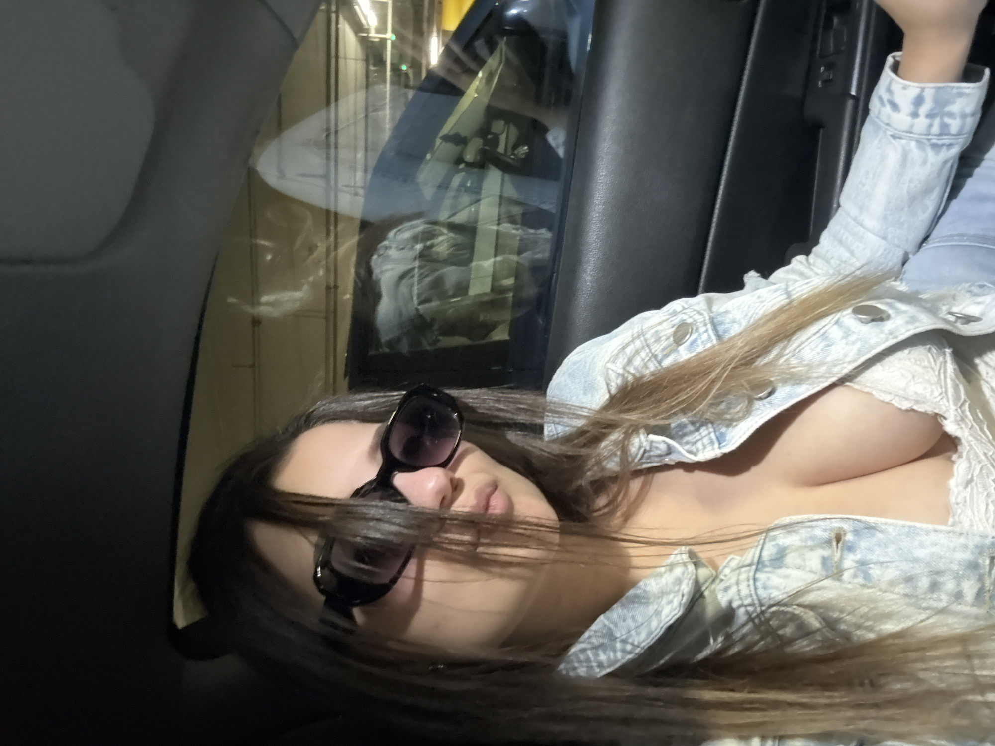 A woman with long brown hair and sunglasses is lying down in a car. She is wearing a light-colored denim jacket over a white top with lace detailing. The car's interior is visible, including the door panel and window. Reflected in the window are blurred lights and shapes that could be urban infrastructure. Without any additional information it's impossible to identify the city and a forensic-matter-of-fact description needs to include this missing information.