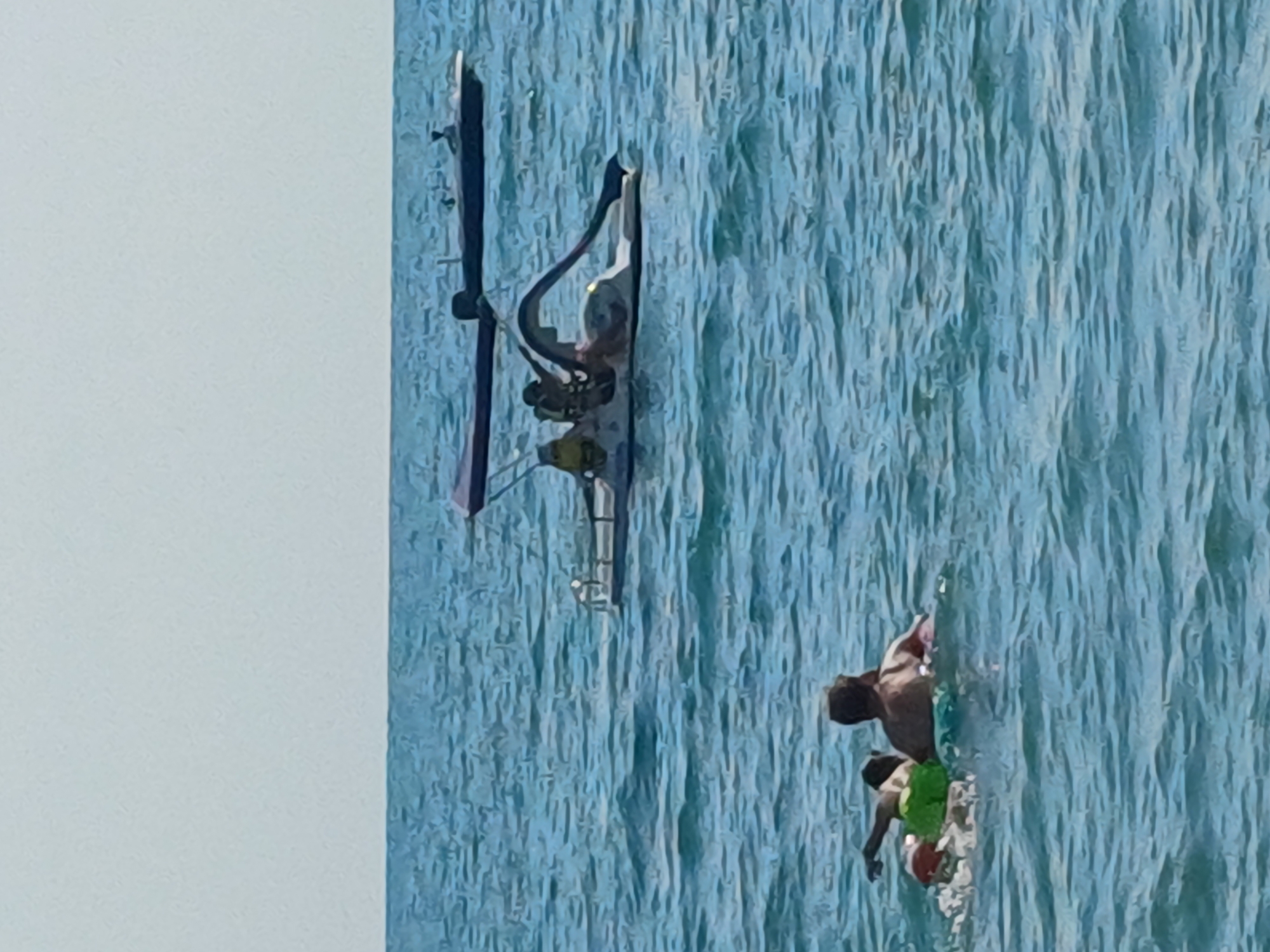 The image shows a person in the water, appearing to be swimming or playing. The water is a vibrant blue and appears to have ripples, suggesting movement or waves. In the background, there is a white or light blue sky. On the left side of the image, there is a dark, upright object that could be a pole or a part of a structure, with some lighter colored elements attached to it, possibly a device or equipment. The lighting suggests it is daytime, with clear skies. The overall impression is of a recreational scene by the water.