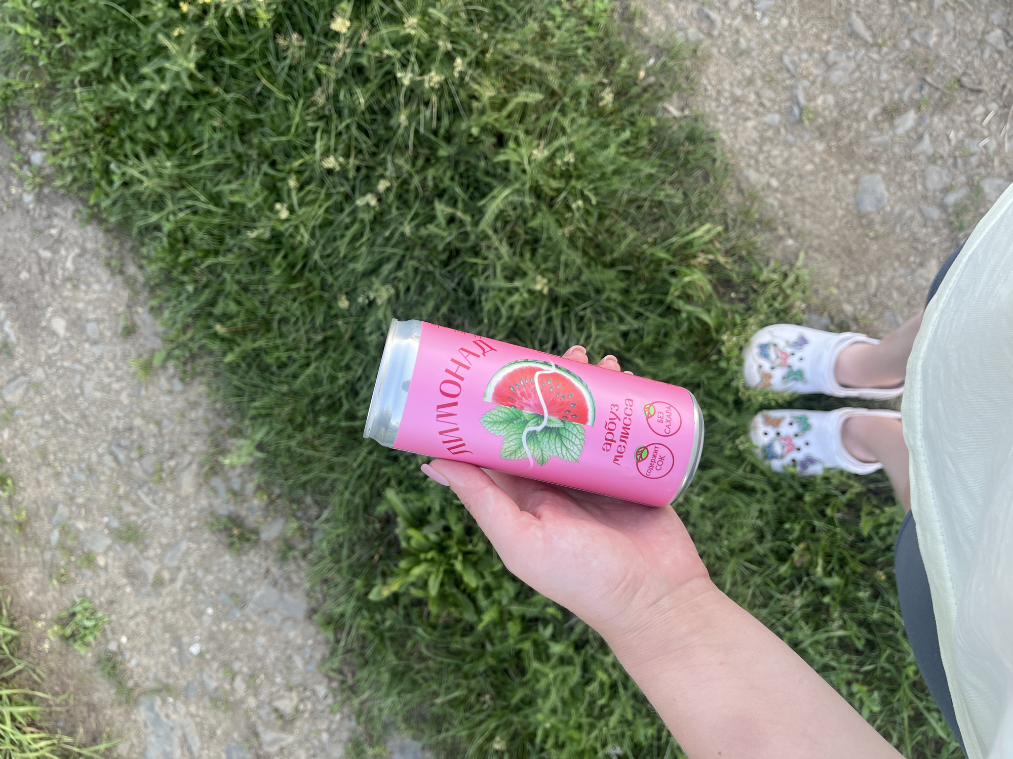 A person's hand holds a pink soda can with a watermelon and mint design. The can has Russian text which appears to say "ЛИМОНАД" on the left side and "арбуз мелисса" (watermelon balm) and "БЕЗ САХАРА" (sugar-free) on the right. The background shows green grass and a gravel path. The person is wearing white clogs decorated with colorful cartoon characters. The scene suggests an outdoor setting, likely during the daytime, given the bright lighting. The context provided places the location in Brusnyatskoye, Russia.