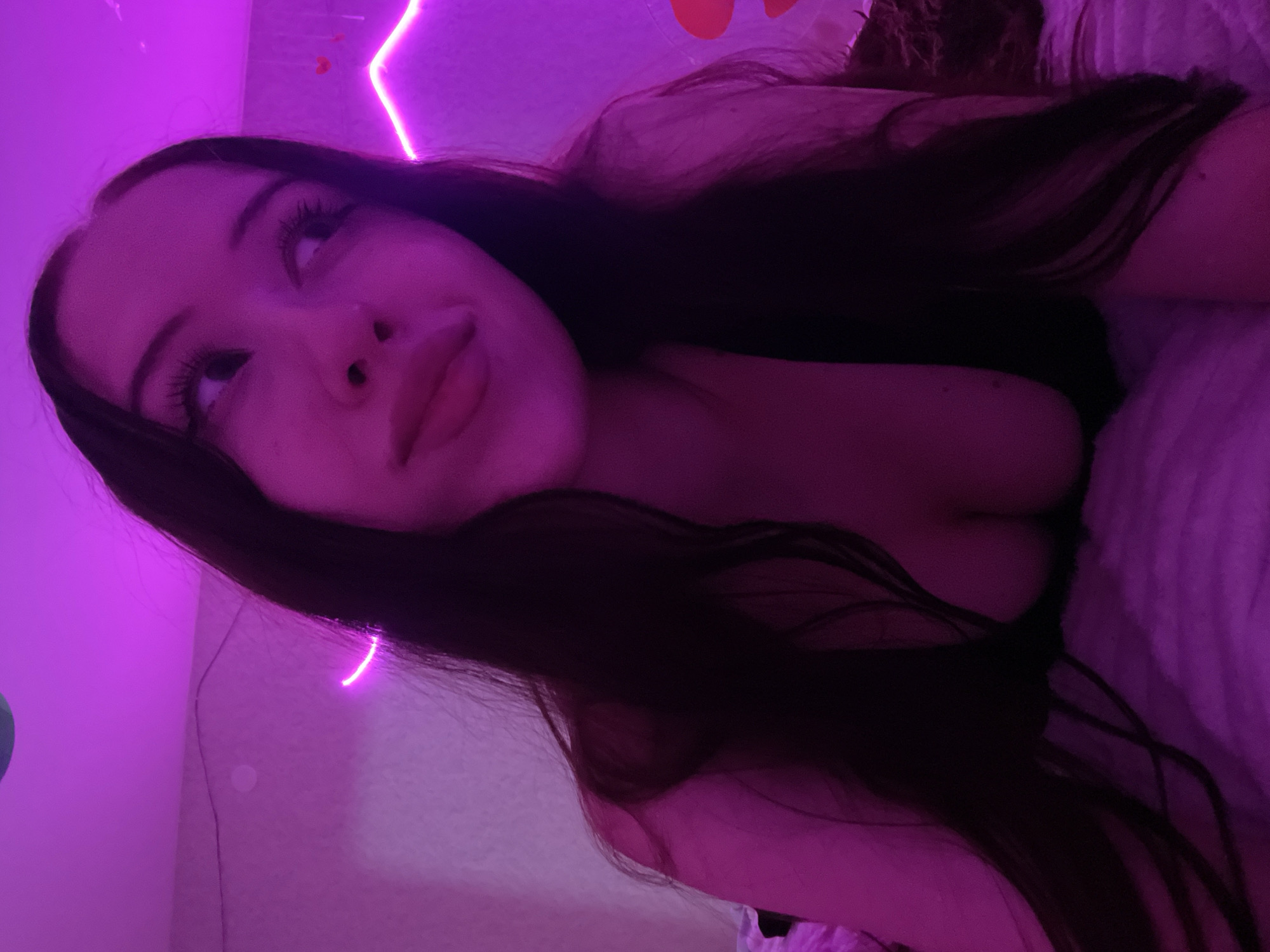 A young woman with long, dark hair lies on a bed, illuminated by purple neon lights. Her eyes are looking to the right, and her lips are slightly parted. She is wearing a dark top that reveals her décolletage. The background consists of a pink wall with neon lighting and some small heart-shaped decorations. The bed is covered with a textured blanket. The scene appears to be an indoor setting, likely a bedroom. There is no information available regarding the location in Votkinsk, Russia.