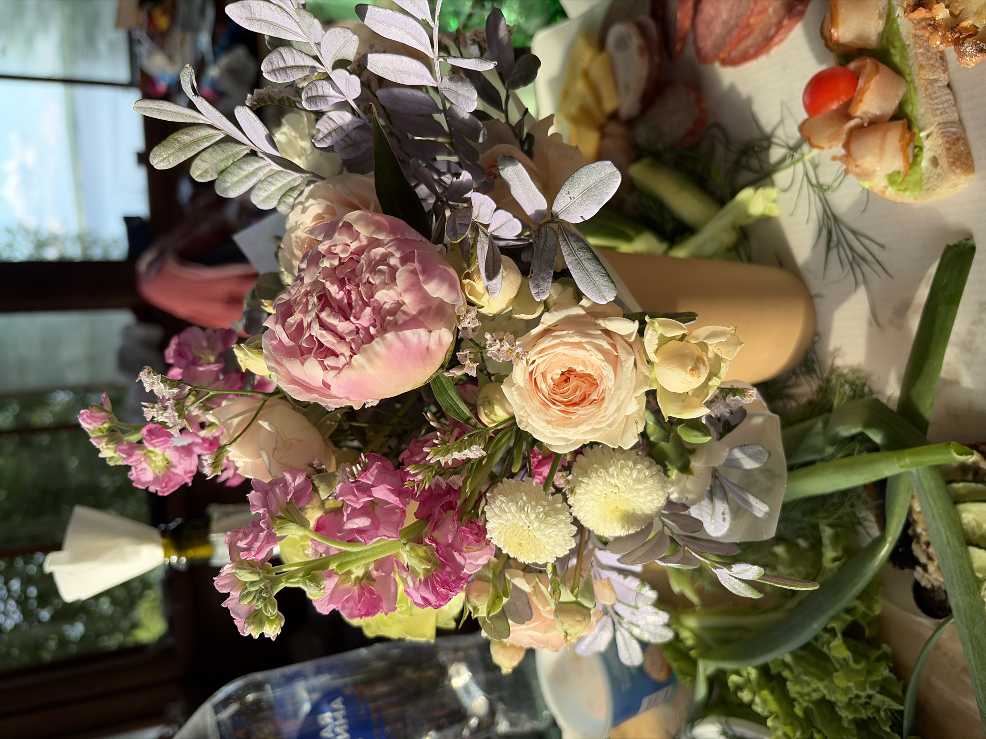 A close-up shot captures a bouquet of flowers, including pink peonies and roses, arranged in a light brown vase. The bouquet is positioned on a table laden with food, featuring various meats, sliced cucumbers, and a small sandwich topped with lettuce, bacon, and a cherry tomato. In the background, a body of water is visible through a window, suggesting an outdoor setting or a room with a view. The lighting indicates it is daytime, with sunlight casting shadows and illuminating the scene. The overall ambiance is one of a relaxed gathering or meal. The presence of water in the background could imply a location near the Kama River, which flows through Chaikovsky, Russia.