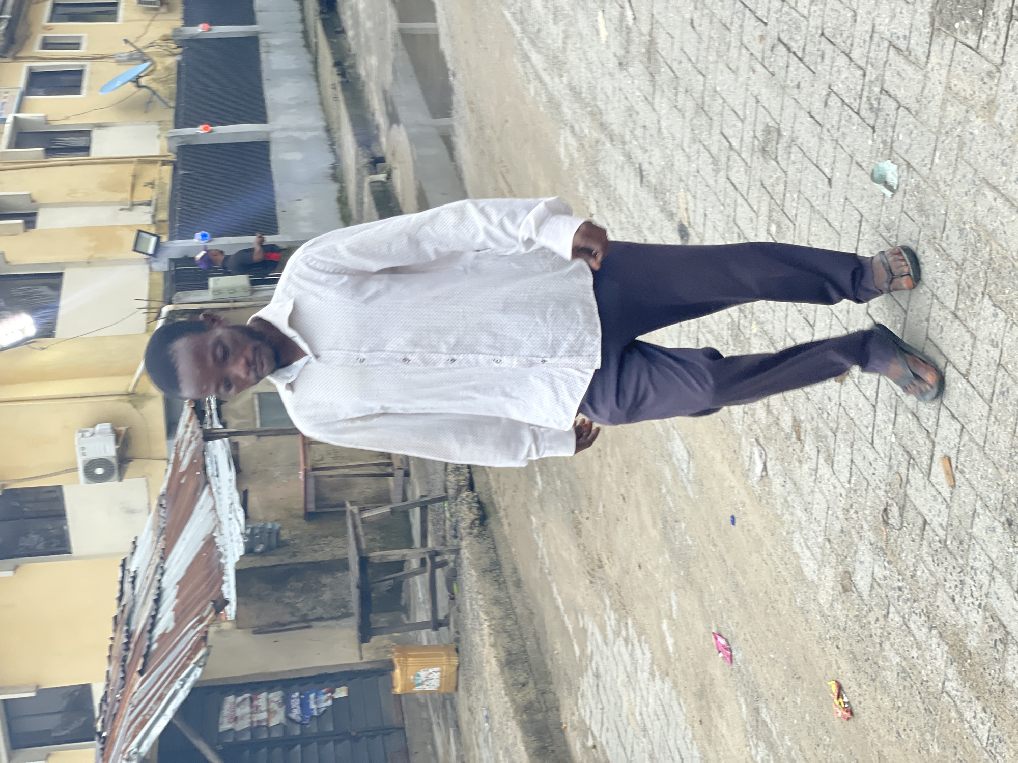The image displays an outdoor urban street scene in Lagos, Nigeria. In the foreground, a dark-skinned male, estimated to be between 20 and 40 years old, walks on a paved surface towards the viewer's left, with his head slightly bowed and gaze directed downwards. He wears a white, long-sleeved collared shirt with a subtle patterned texture, dark-colored trousers, and dark-colored flip-flops. His right arm is bent at the elbow, while his left arm hangs alongside his body.
The ground is paved with interlocking concrete blocks, but it is largely obscured by a layer of dirt and sand, exhibiting damp patches or small puddles. Scattered litter includes pink and yellow plastic wrappers near the bottom left, a small blue object in the center, and a crumpled white and green item near the main subject's right foot.
To the left of the main subject in the midground, a single-story structure features a heavily rusted corrugated metal roof and rough, light-colored walls. A dark shuttered opening on this structure is adorned with multiple posters or flyers. A simple rectangular wooden table stands on the ground in front of this structure, with a yellow plastic jerry can positioned nearby.
Further into the background and to the right, a multi-story building, at least two stories high, is painted a light yellow and features multiple dark-paned windows. An outdoor air conditioning unit and a small blue satellite dish are mounted on this building's exterior. A dark metal fence, interspersed with concrete pillars, runs in front of the yellow building. Red and blue lights are attached to these fence pillars. A second, partially obscured dark-skinned male, wearing a dark shirt and a purple cap, stands near this fence, positioned between the main subject and the yellow building. The overall lighting is diffused, indicating an overcast sky or a time of day with indirect sunlight.