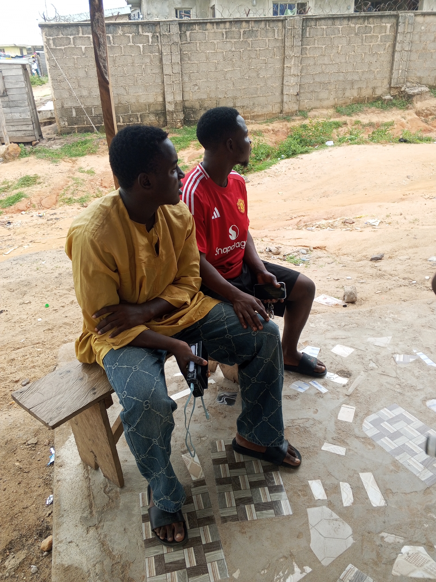 Two men are sitting on a wooden bench outside a building in Lokoja, Nigeria. The man on the left is wearing a yellow traditional African shirt and blue jeans with a repeating pattern. He is holding a black object, possibly a phone or camera. The man on the right is wearing a red Manchester United jersey and black shorts. He is looking to his right and holding a black phone in his right hand. The ground is a mix of sand and patterned tiles. In the background, there is a brick wall with barbed wire on top, and a building with windows. To the left of the bench, there is a wooden structure and more sandy ground with some dry grass. The overall setting appears to be an outdoor area associated with a residential or commercial building.