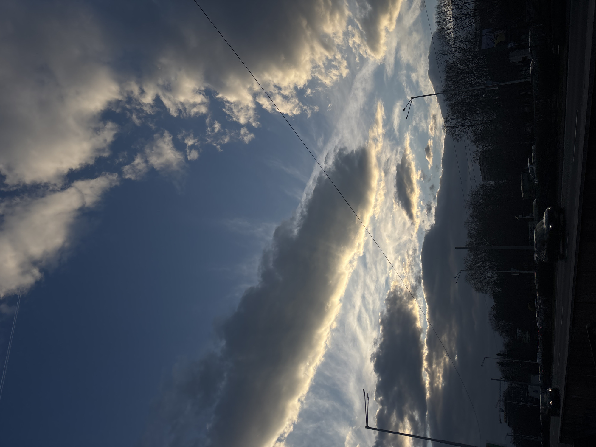 The image displays an urban street scene in Sofia, Bulgaria, observed during dusk or late afternoon. The sky dominates the upper two-thirds of the frame, characterized by a deep blue interspersed with large, dark gray clouds. Direct sunlight illuminates the lower edges of these clouds from behind, creating prominent golden and bright white highlights. Wispy white cirrus clouds are visible in various parts of the blue sky, along with a faint aircraft contrail in the upper left quadrant. Multiple dark overhead power lines or trolley wires traverse the sky, notably from the lower right to the upper left.
The lower third of the image features a multi-lane road, partially obscured by a dark concrete barrier in the immediate foreground. Several vehicles, predominantly dark passenger cars, are visible on the road, silhouetted against the brighter sky with illuminated headlights. Two tall, dark streetlights with double arms are positioned on either side of the road. Behind the road, a dense line of bare, deciduous trees forms a dark, silhouetted barrier across the mid-ground. Beyond the trees, the faint outlines of urban buildings are visible, including a construction site with a crane on the far left. The scene is characterized by low ambient light conditions.