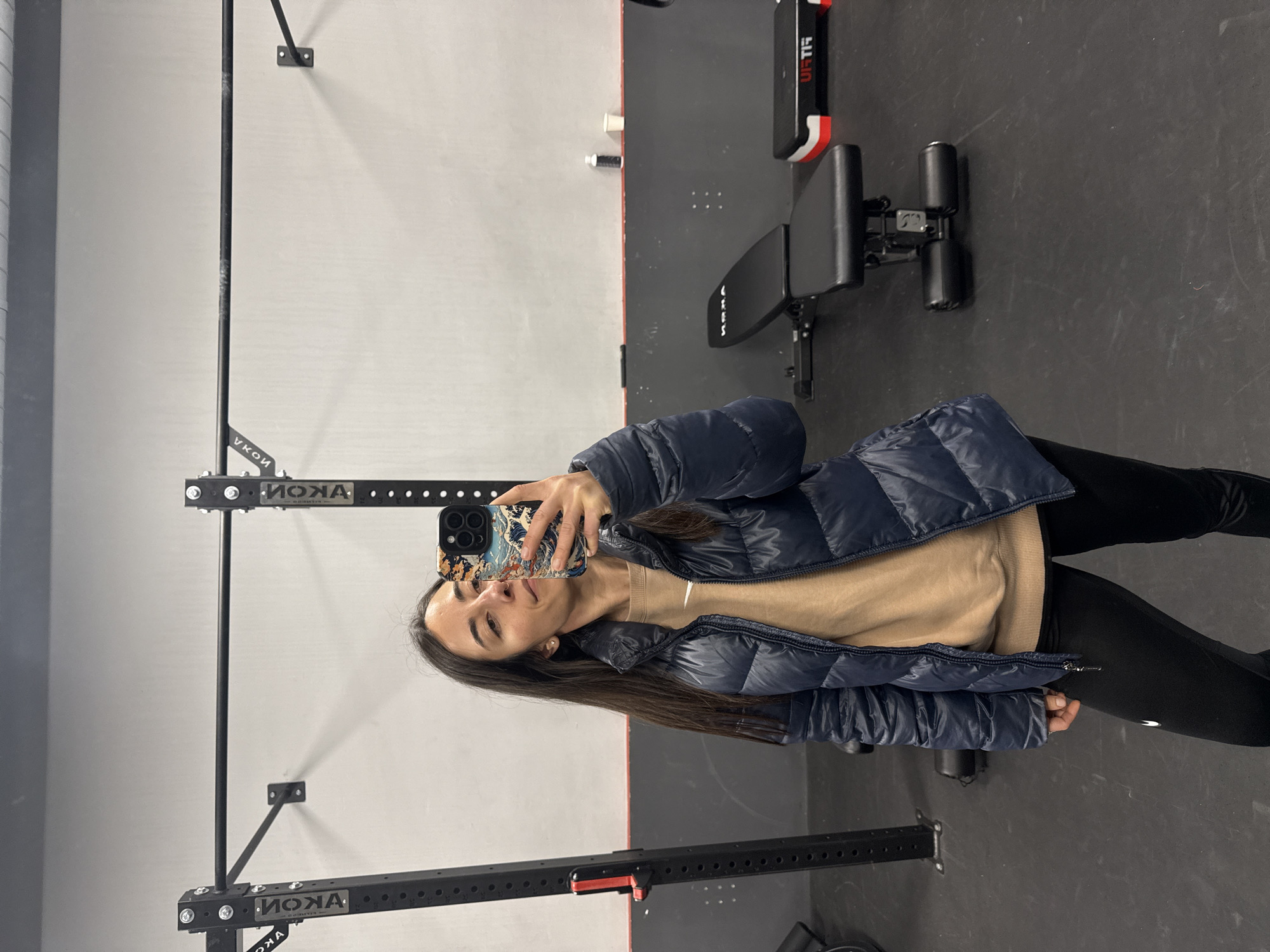 The image is oriented vertically, rotated 90 degrees counter-clockwise, and depicts a young woman taking a mirror selfie inside a gym. She has long, straight dark brown hair and a neutral expression, possibly with a subtle smile. She is dressed in a dark blue puffer jacket over a light beige long-sleeved top and black leggings. She holds a smartphone with her left hand, which is adorned with a distinctive phone case featuring an intricate design reminiscent of Japanese ukiyo-e art, specifically "The Great Wave off Kanagawa."
The setting is a contemporary fitness facility, likely located in Barcelona, Spain. The background consists of light grey walls and a dark grey floor. Various pieces of black gym equipment are visible. On the left side of the frame, a black power rack or squat rack is prominent, displaying the brand name "AKON" in white capital letters on its structure. Towards the right, a black adjustable weight bench also features the "AKON" logo on its upholstery. Another piece of exercise equipment, possibly a sissy squat bench with black padded rollers, is partially visible on the far right. Red and white trim on some background equipment shows the brand "VIRTIS." The overall environment suggests a functional and well-equipped workout space, captured under bright indoor lighting. The activity is a casual self-portrait taken within this fitness environment.