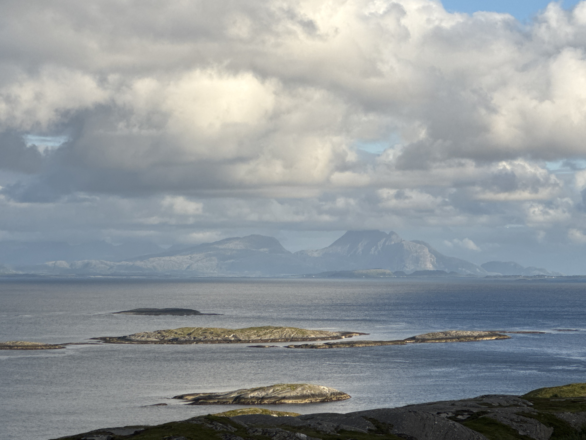 This is a wide, horizontal shot of the Norwegian coastline near Sleneset, Norway. The foreground shows dark, rocky terrain covered with sparse green vegetation. The middle ground is dominated by calm, blue-grey water with several small, rocky islands dotting its surface. These islands vary in size and shape, with some appearing barren and others having patches of green grass. In the background, a distant mountain range rises, its peaks shrouded in a hazy, atmospheric perspective. The sky above is filled with large, dramatic clouds, some white and illuminated by sunlight, others darker and grey, suggesting an overcast or partly cloudy day. There are no people or animals visible in the image, and no discernible actions or interactions are taking place. The overall scene is one of natural, rugged beauty, with a sense of vastness and tranquility.