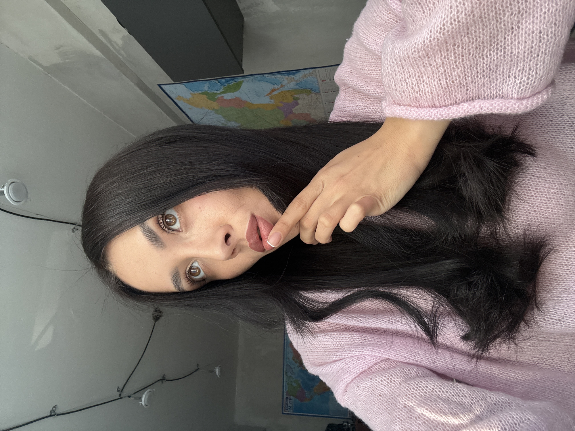 A woman with long, dark hair and fair skin is wearing a light pink sweater. She is holding her finger to her lips. The background includes a wall, a world map, and string lights. It is not possible to determine from the image whether the scene is in Votkinsk, Russia.