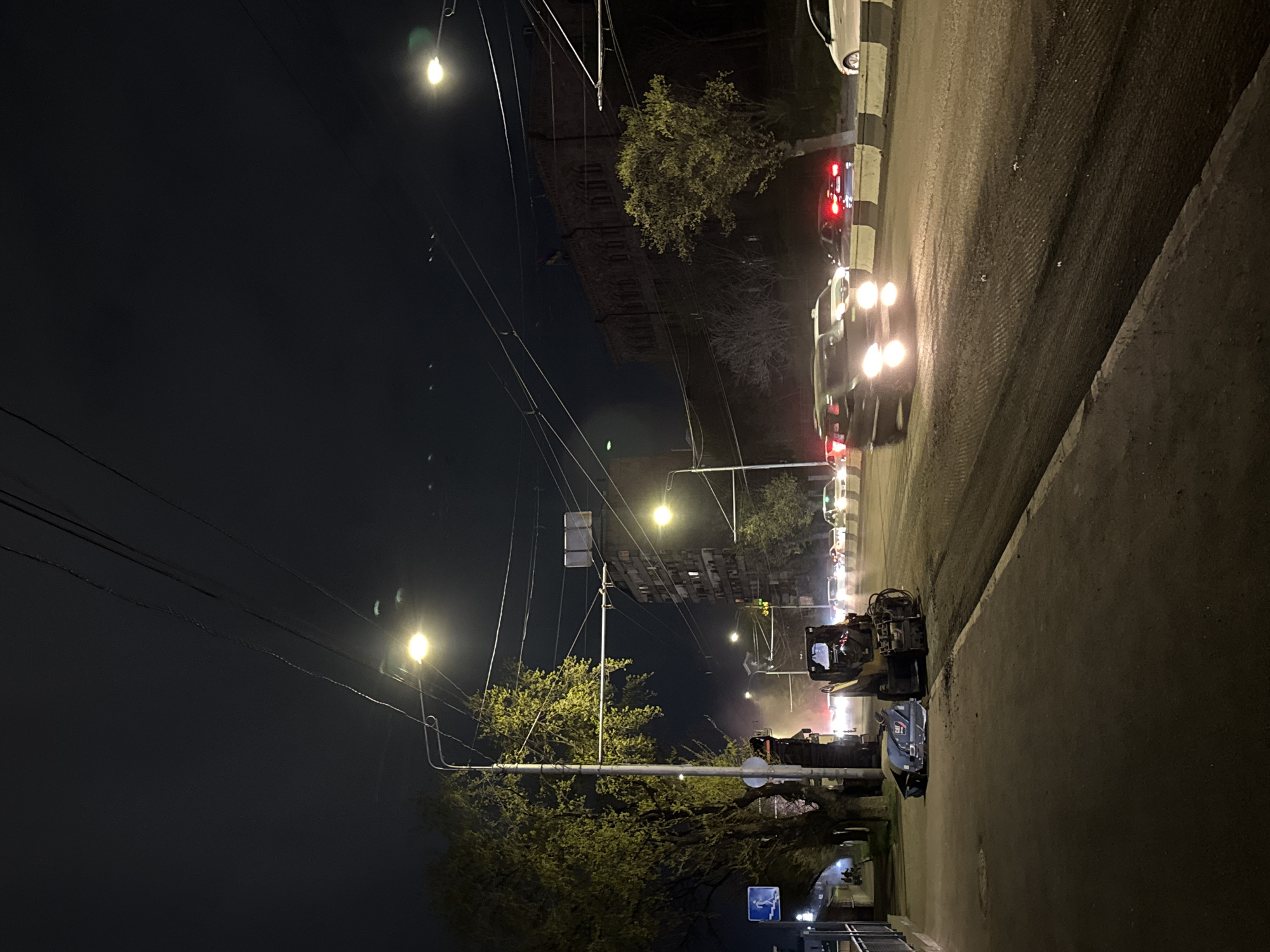 At night in Yerevan, Armenia, a street scene shows road repair operations. A road milling machine sits on the right edge of the road; in front of it is a car with a small overturned trailer. In the background, traffic moves through the city. Street lights illuminate the scene. Several buildings stand along the street, including a large building in the upper left. Power lines cross the night sky.