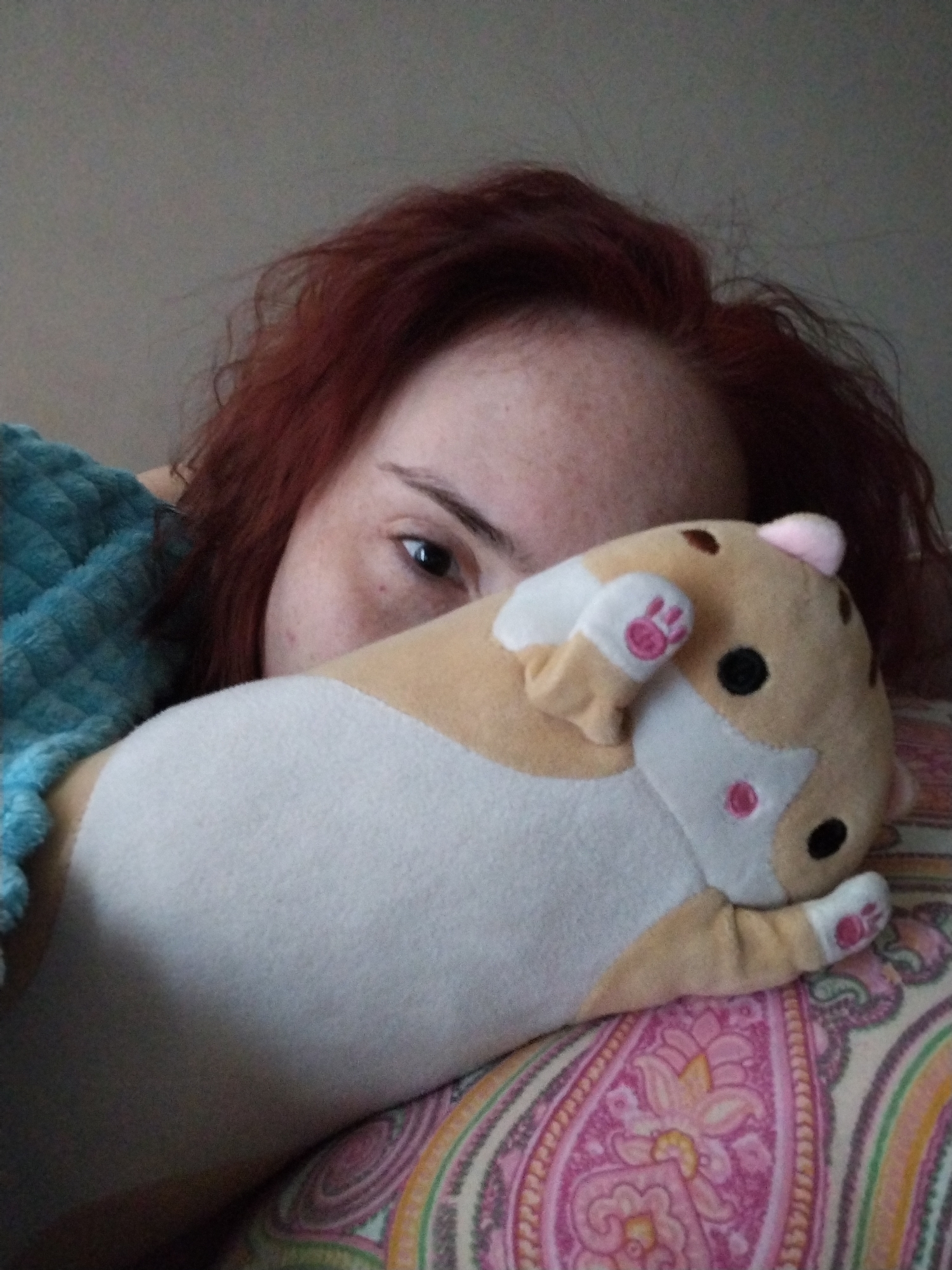 A woman with red hair is lying down, partially visible, with a stuffed animal resting on a patterned pillow. She is partially covered by a blue, textured blanket. Only one eye is fully visible. The location is not discernible as Izhevsk, Russia.