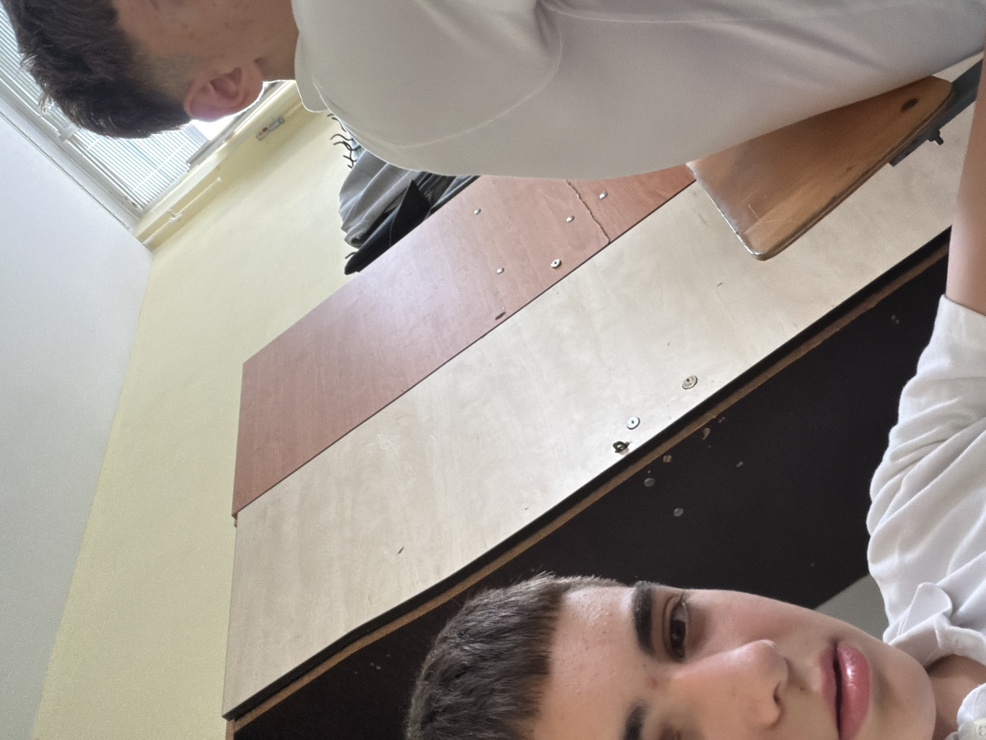 Two individuals are captured in this close-up, angled shot. Both are wearing white collared shirts. One person, positioned at the top of the frame, shows only a portion of their head and shoulder. The other person, located at the bottom right of the frame, displays their face more prominently, including their eyes, nose, and mouth. The setting appears to be indoors, possibly a classroom or similar academic environment, with wooden desks visible. The location is Varna, Bulgaria. Details of the surroundings include a wall, a window with blinds, and other obscured objects, possibly clothing or bags, which are only partially visible. The interaction between the people or their specific actions is not clear from the image.