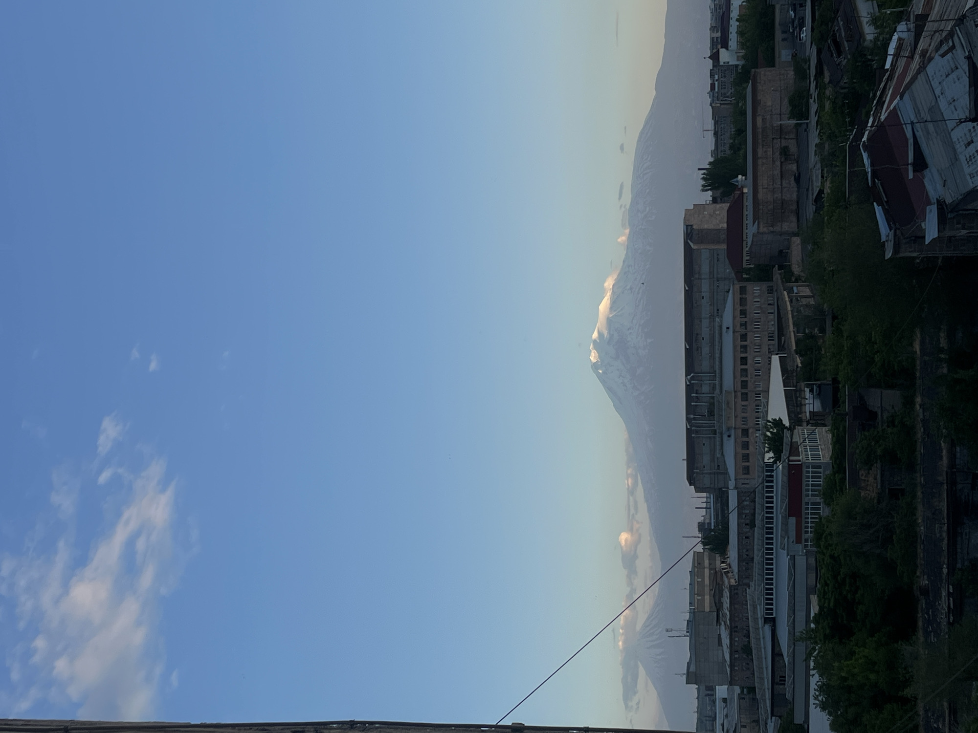 The image shows a view of Mount Ararat from Yerevan, Armenia. The left side of the image is dominated by a clear blue sky with some wispy clouds. On the right side, there's a cityscape with buildings of varying heights and architectural styles. Some buildings appear older and more dilapidated with visible decay, while others seem more modern. Patches of greenery indicate trees interspersed among the buildings. The image is taken from a slightly elevated vantage point, possibly from a rooftop or upper floor. A wire cuts diagonally across the lower central part of the frame.