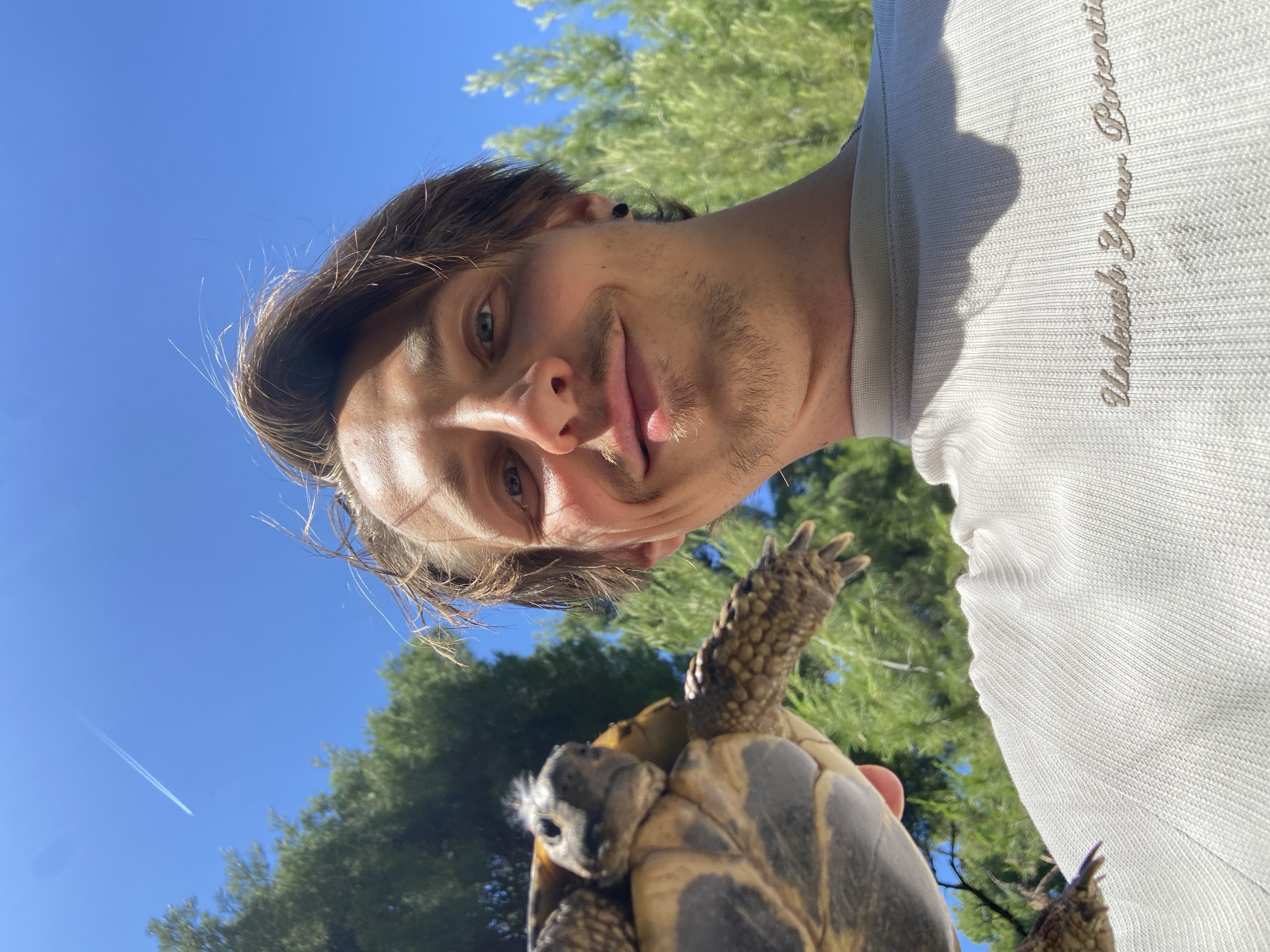A low-angle selfie taken outdoors in daylight. A man with light brown hair and stubble is holding a tortoise. The man wears a light gray shirt. The sky is blue, and contrails from an airplane are visible in the background. Green trees are present in the background as well. The location is likely Šušanj, Montenegro, considering it is in the title of the image.