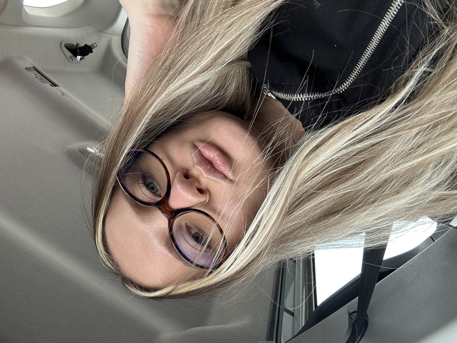 The scene depicts a female individual taking a selfie inside a vehicle. The given context places this vehicle in Torbalı, Turkey. The subject is a light-skinned woman with long, straight blonde hair featuring darker undertones or highlights, extending over her shoulders and chest. She wears brown tortoise-shell framed eyeglasses, and a small silver hoop nose ring is visible on her left nostril. Her lips are slightly pursed in an expressive pose. She is dressed in a black garment, possibly a jacket or hoodie, characterized by a prominent silver zipper running vertically on the right side of the frame. Her left hand is partially visible at the top right of the frame, with fingers touching her hair, suggesting she is holding the camera or phone used for the photograph. The background consists of the light grey or off-white interior headliner of a vehicle, with an overhead fixture or cutout visible. A dark strap or handle, possibly a seatbelt component or grab handle, is also visible on the upper right side of the vehicle interior. A window frame is partially visible on the far left, revealing an undifferentiated bright exterior.