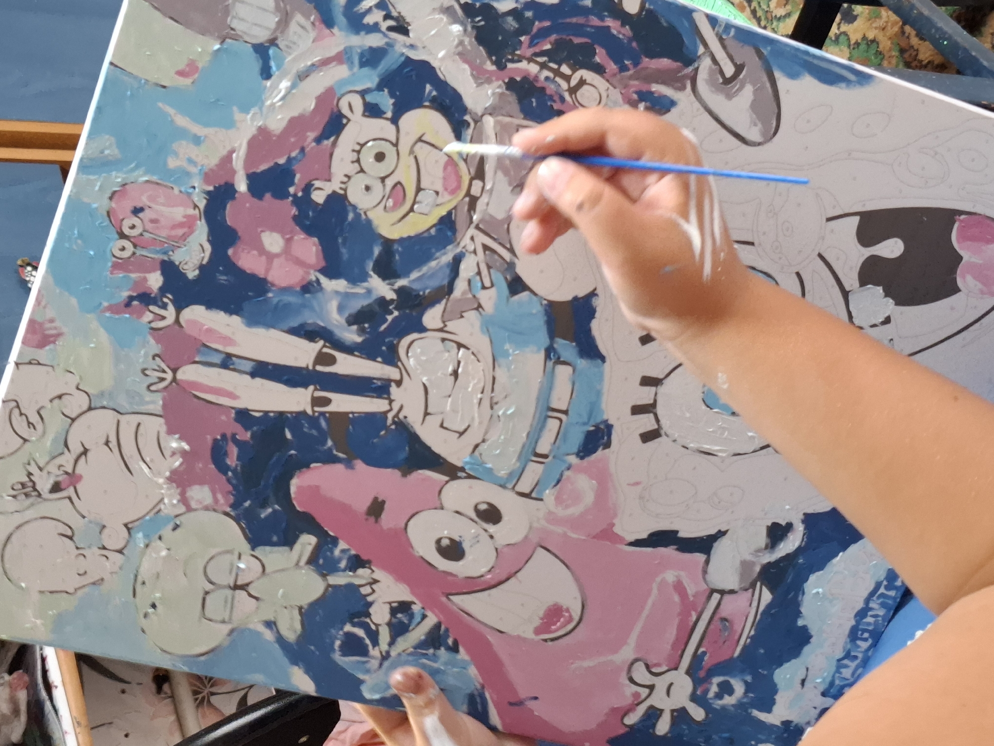 The image shows a person's hands painting a canvas featuring characters from SpongeBob SquarePants. The canvas is partially painted with vibrant colors, including blue, pink, and white, and the outlines of SpongeBob, Patrick, Squidward, Sandy, and Mr. Krabs are visible. A blue paintbrush is being used to add paint to the canvas. The scene is lit by natural light and appears to be an indoor setting, possibly a home. The background is blurred, but suggests a shelf with items and a wooden support structure. The location is not specified in the image or context provided.