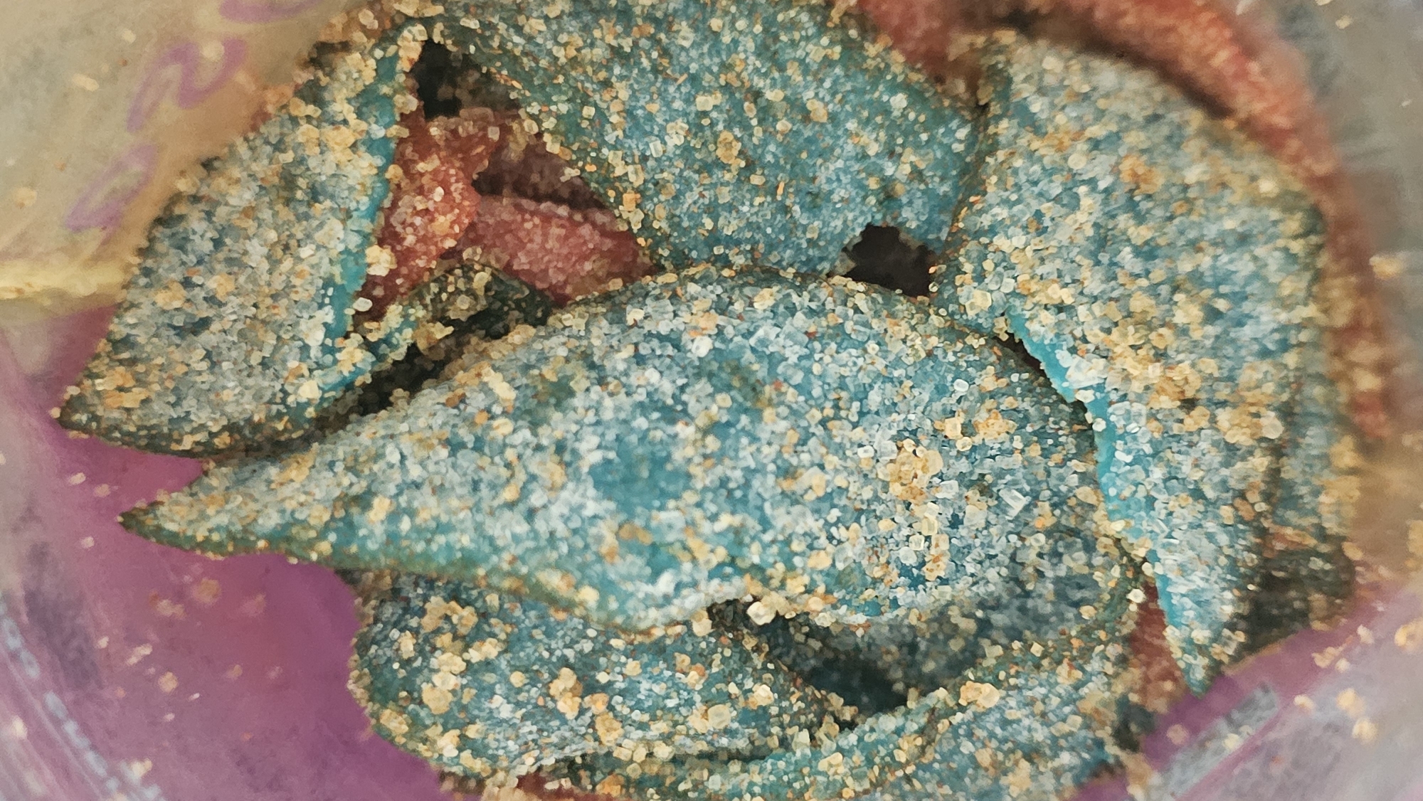 A close-up shot depicts a pile of blue and red gummy candies, heavily coated in granulated sugar. The candies appear to be in a bag, possibly plastic, with hints of pink and clear plastic visible around the edges. The location details are not applicable to this image.