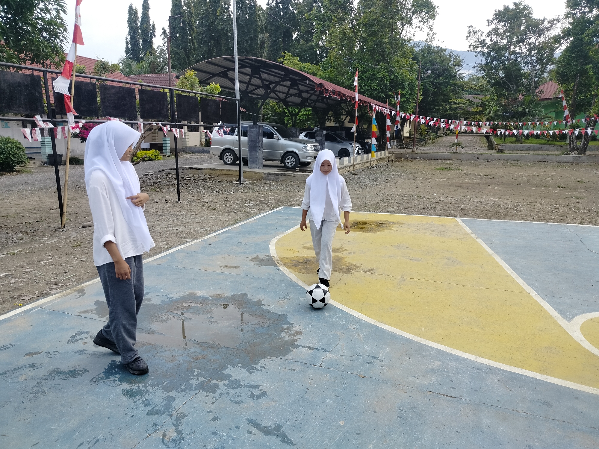 Two young girls wearing white hijabs and modest clothing are on a wet outdoor basketball court in Lawebekung, Indonesia. The girl on the left, facing away from the camera, wears a white long-sleeved shirt and grey pants. She is standing on the blue portion of the court. The girl on the right, facing the camera and slightly to the left, wears a similar white long-sleeved shirt and grey pants. She is standing near the center of the court, between the yellow and blue painted sections, and is actively kicking a black and white soccer ball with her right foot. Puddles of water are visible on the blue painted surface of the court. In the background, there is a covered parking area with several silver cars parked underneath. Red and white Indonesian flags are visible, some attached to poles and others strung as pennants across the area. Trees line the perimeter of the scene, with buildings and possibly a mountain range visible in the distance under an overcast sky. The environment appears to be an outdoor recreational area, possibly associated with a school or community center.