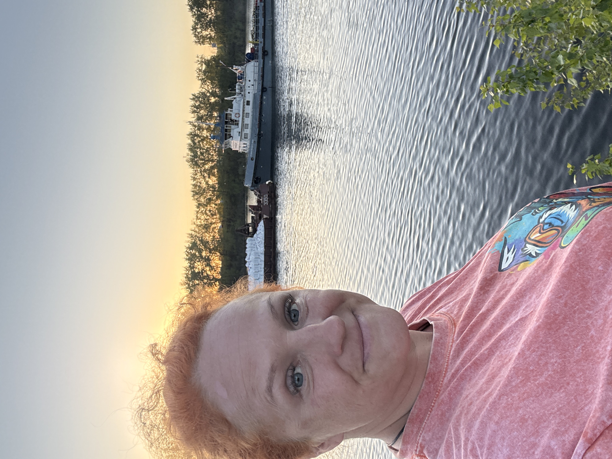 A woman with red hair is standing outside, likely at sunset, with a body of water and a boat visible in the background. The body of water is possibly the Kama River. The boat appears to be a barge or workboat, which, along with the river, suggests a possible location in Chaikovsky, Russia. The environment includes trees and foliage along the riverbank. The woman is wearing a pink shirt with a graphic design.