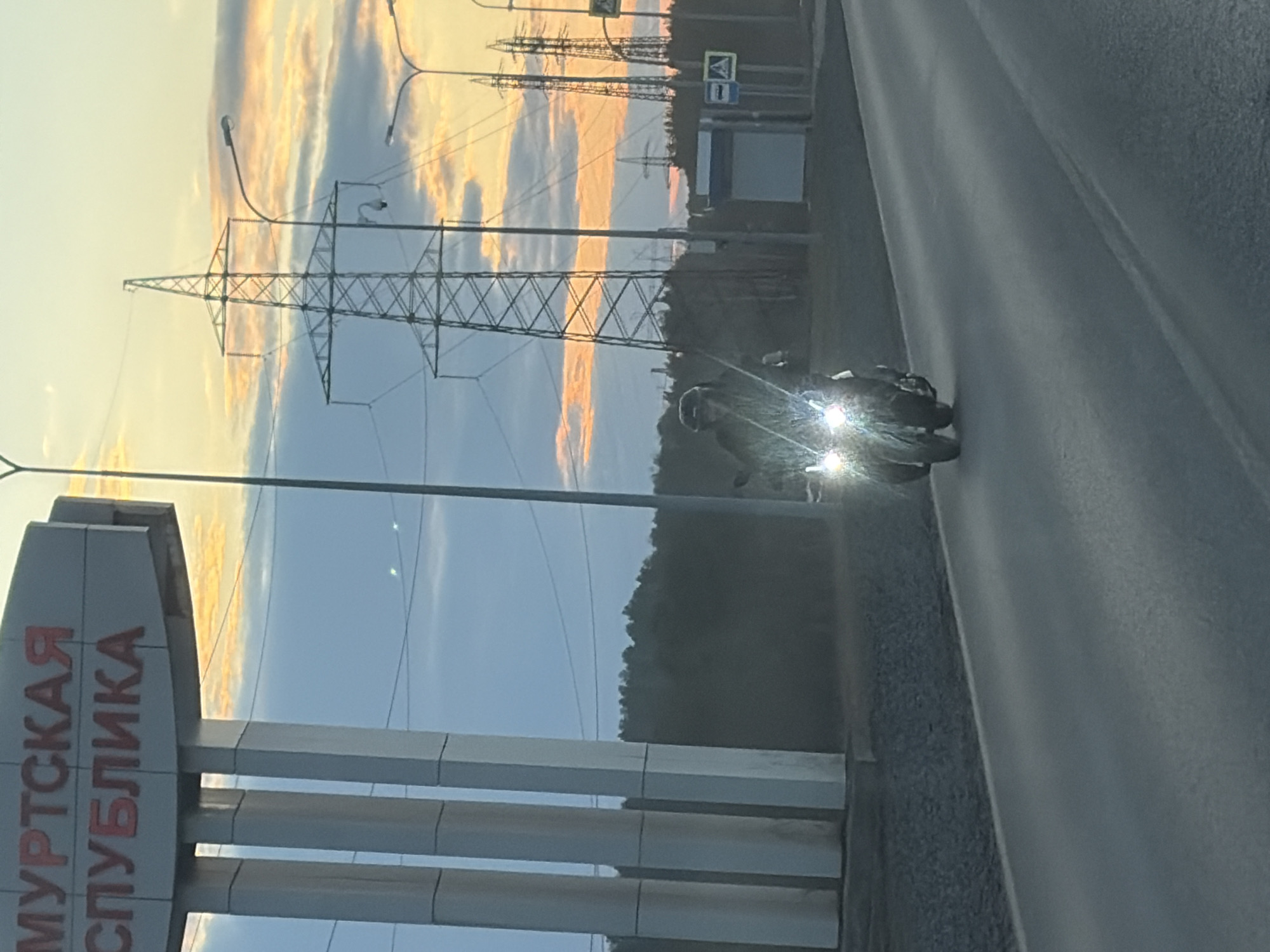 The image shows a road with a motorcycle driving on the right side, towards the viewer. Power lines and street lights are visible in the background, along with a sign that reads "Удмуртская Республика" in red letters. The sky is visible above the power lines and features a sunset-like glow with shades of orange and blue. A forest is present in the background, adding to the natural setting of the shot. The road appears to be in Chaikovsky, Russia.