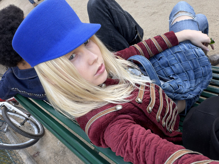 The image depicts three individuals seated on a green slatted park bench within a public garden in Paris, France. The central subject, a person with long blonde hair featuring a subtle red streak, wears a bright blue felt brimmed hat and a dark red quilted jacket adorned with gold-colored braided trim and silver buttons. This individual is looking directly at the camera. To their left, a person with dark, curly hair is partially visible from the back, wearing a dark denim jacket with studded embellishments. To the right, the lower body of a third individual is visible; they wear light blue ripped jeans and dark trousers, and a brown shoe is on the ground beside the bench. This person's right hand, wearing a green ring, holds a small green cylindrical object. A woven light blue denim bag rests on the lap of the individual in ripped jeans. A white bicycle is partially visible on the ground to the far left, adjacent to the bench, with a chain-link fence behind it. The ground surface is light brown, consistent with sand or fine gravel.
