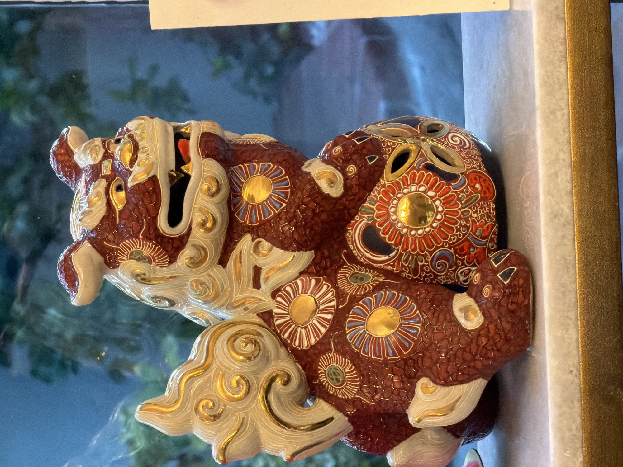 A detailed, ceramic statue of a Foo dog, also known as a guardian lion, is positioned on a white marble shelf with a golden trim. The Foo dog is predominantly a deep red color, with intricate patterns of gold and white swirling designs adorning its mane, back, and tail. It features a fierce expression with its mouth open, revealing a red tongue, and its eyes are a bright yellow. The statue is decorated with circular motifs of blue, red, and gold, resembling stylized flowers. The background consists of a blurred blue-toned window, reflecting greenery and possibly a building exterior, suggesting an indoor setting in San Marino, California, United States of America. The lighting is somewhat dim, with a slight glare on the surface of the statue and shelf.