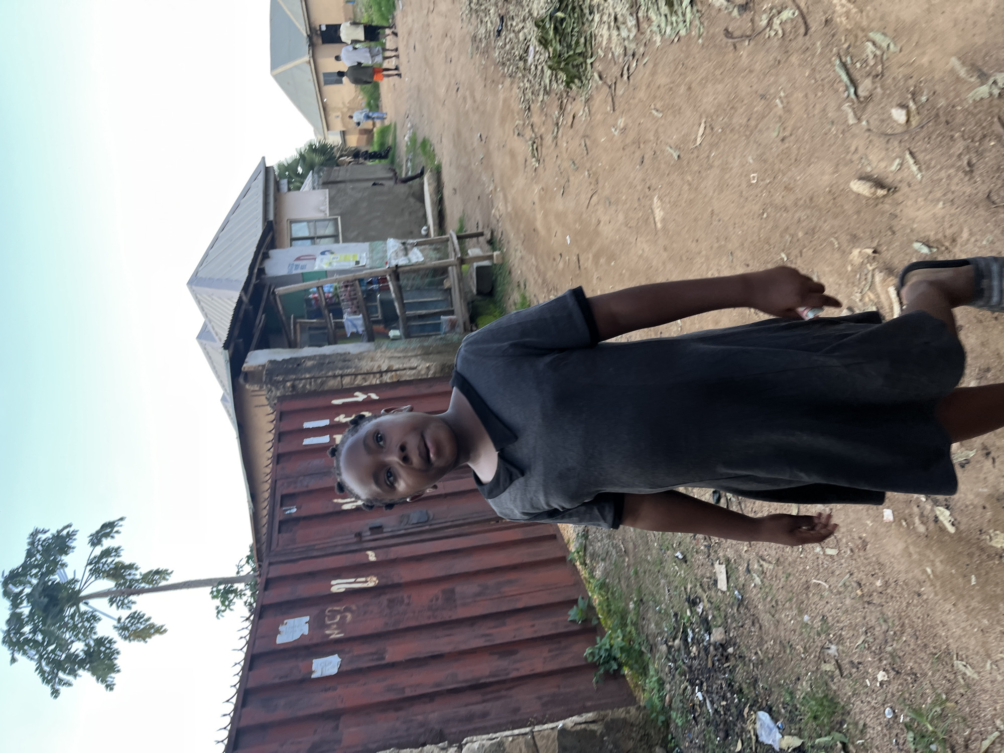 A medium shot shows a young Black girl with her hair in braids, wearing a dark gray dress with a white collar, standing on a dirt path in what appears to be a rural or semi-urban setting in Rafin Par, Nigeria. She is looking to her right, her mouth slightly open. Behind her, to the left, is a tall, reddish-brown corrugated metal fence with some markings on it. To the right of the fence is a small open-air stall or shop with shelves displaying various items. Further in the background, there are buildings with metal roofs and a few people walking away from the camera down the path. The ground is a reddish-brown dirt, littered with dry leaves and some debris. A tall tree with green foliage is visible behind the fence on the left. The sky is clear and pale blue.