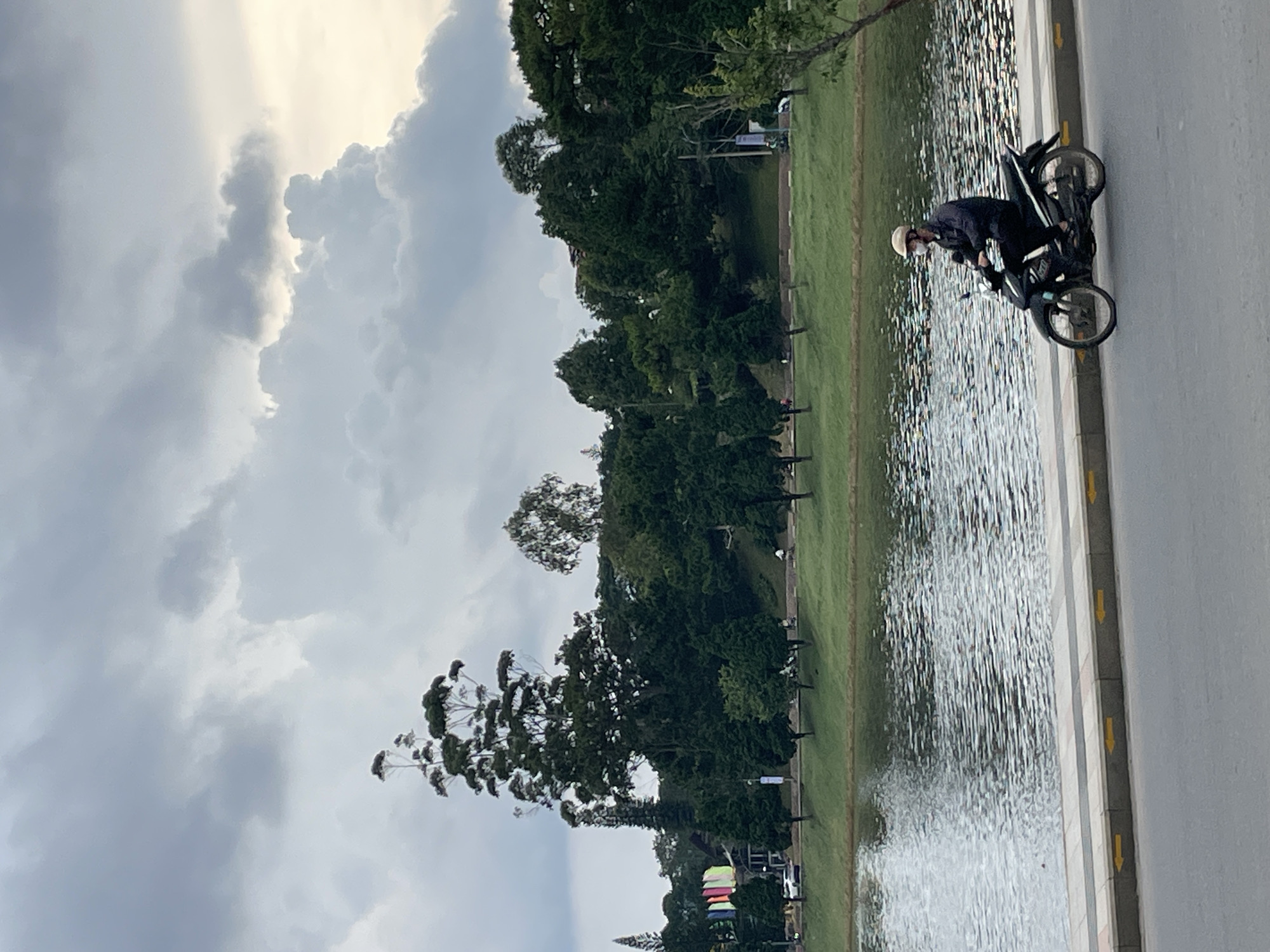A person rides a dark-colored motorcycle on a paved road next to a body of water. The rider wears a light-colored helmet and a face mask. The road has yellow arrows painted on it, indicating direction. In the background, a grassy slope leads up to a dense line of trees. Beyond the trees, a cloudy sky with dramatic cloud formations is visible. The scene appears to be a park or lakeside area in Lâm Viên - Đà Lạt, Vietnam.
