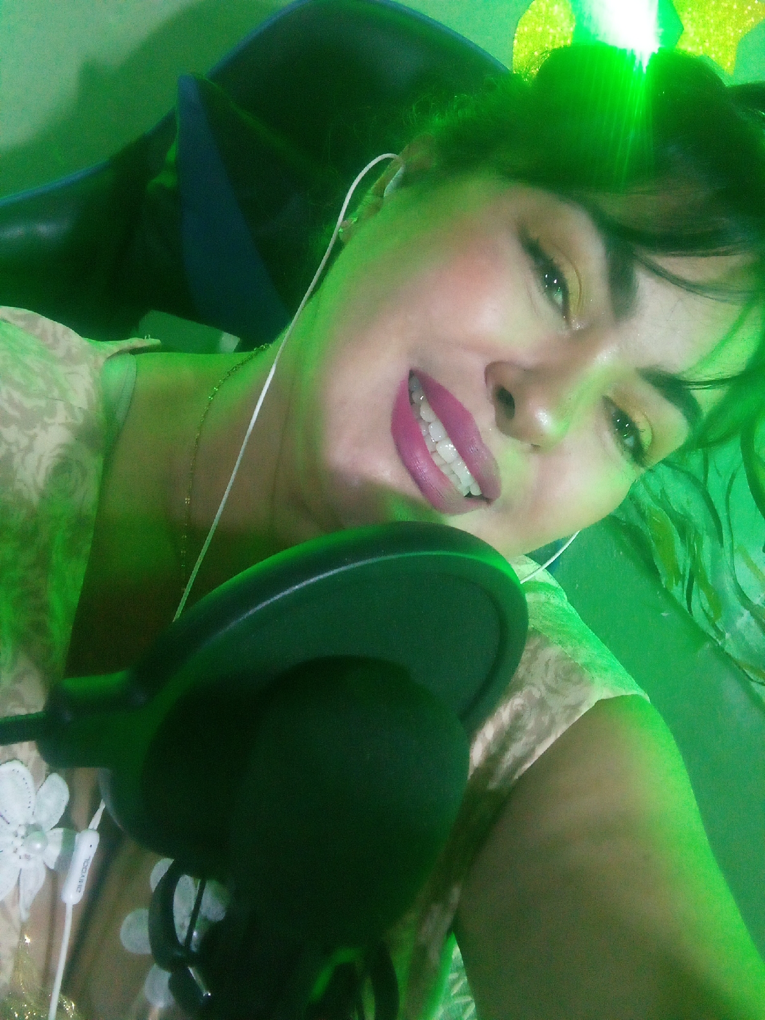 A woman with dark hair and fair skin is shown from the shoulders up, slightly tilted to the left. She is wearing a light-colored patterned shirt, a thin gold necklace, and in-ear headphones. She has pink lipstick and visible makeup. A dark-colored microphone and its stand are positioned in the foreground. She is sitting in a dark chair. The lighting in the scene is predominantly green. The location is unknown.
