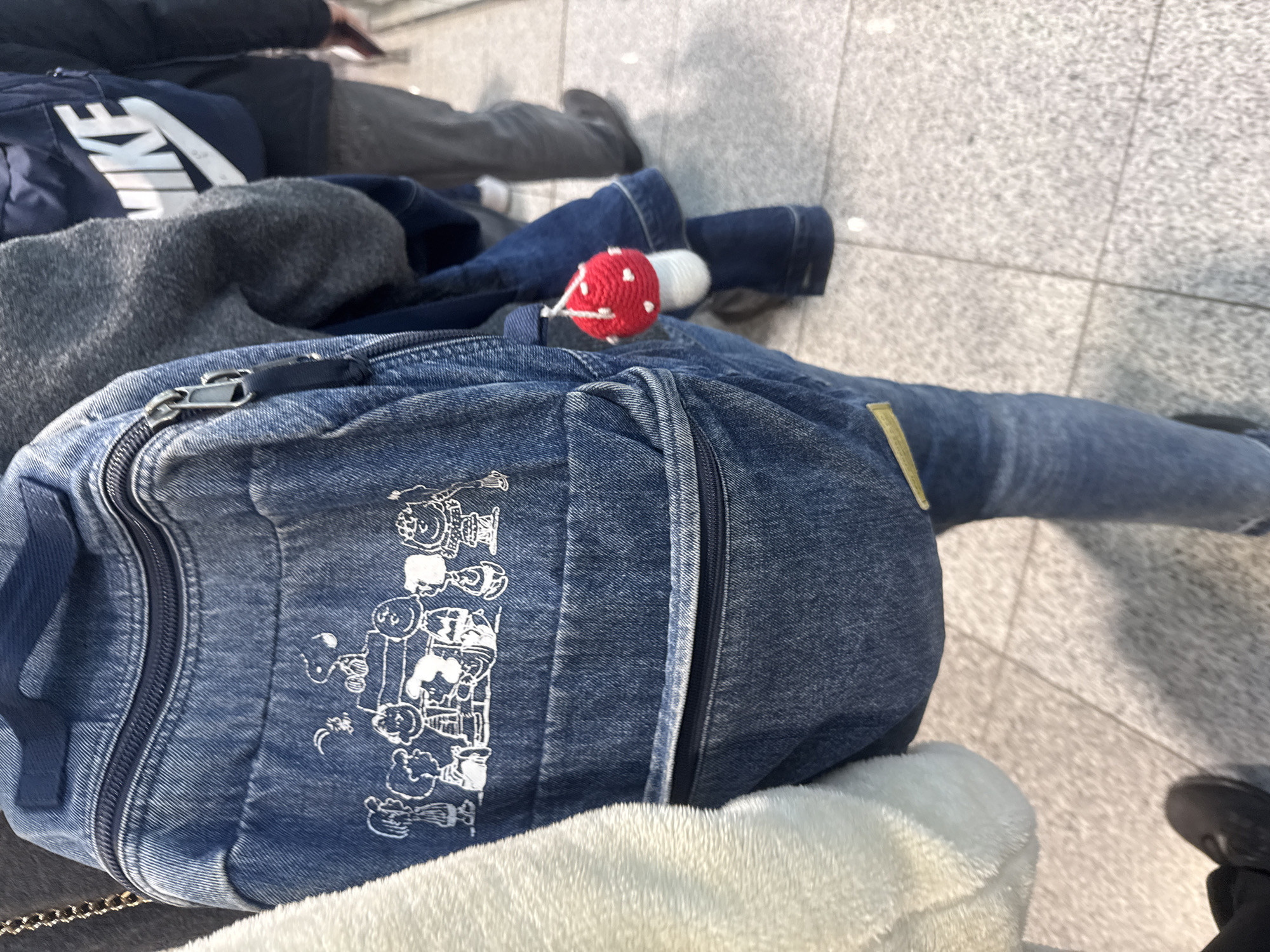 The image depicts an indoor scene in Khimki, Russia, showing a line of at least three individuals from a slightly elevated, rear perspective. In the immediate foreground and center, a person is visible wearing a medium-wash denim backpack. The backpack features a top zipper closure and a lower horizontal zipper pocket. Across the main panel, white outline illustrations of numerous "Peanuts" comic strip characters are depicted. A small, red and white knitted mushroom charm hangs from a loop on the right side of this backpack. The individual is also wearing blue jeans and a grey long-sleeved garment, partially obscured on the left by a white, fluffy garment belonging to an adjacent, mostly unseen person. Further back and to the right, another individual wears a dark blue backpack with a prominent white "NIKE" logo. A third person is partially visible behind them. All individuals are standing on a light grey tiled floor with visible grout lines and a speckled texture, characteristic of a public building's interior.