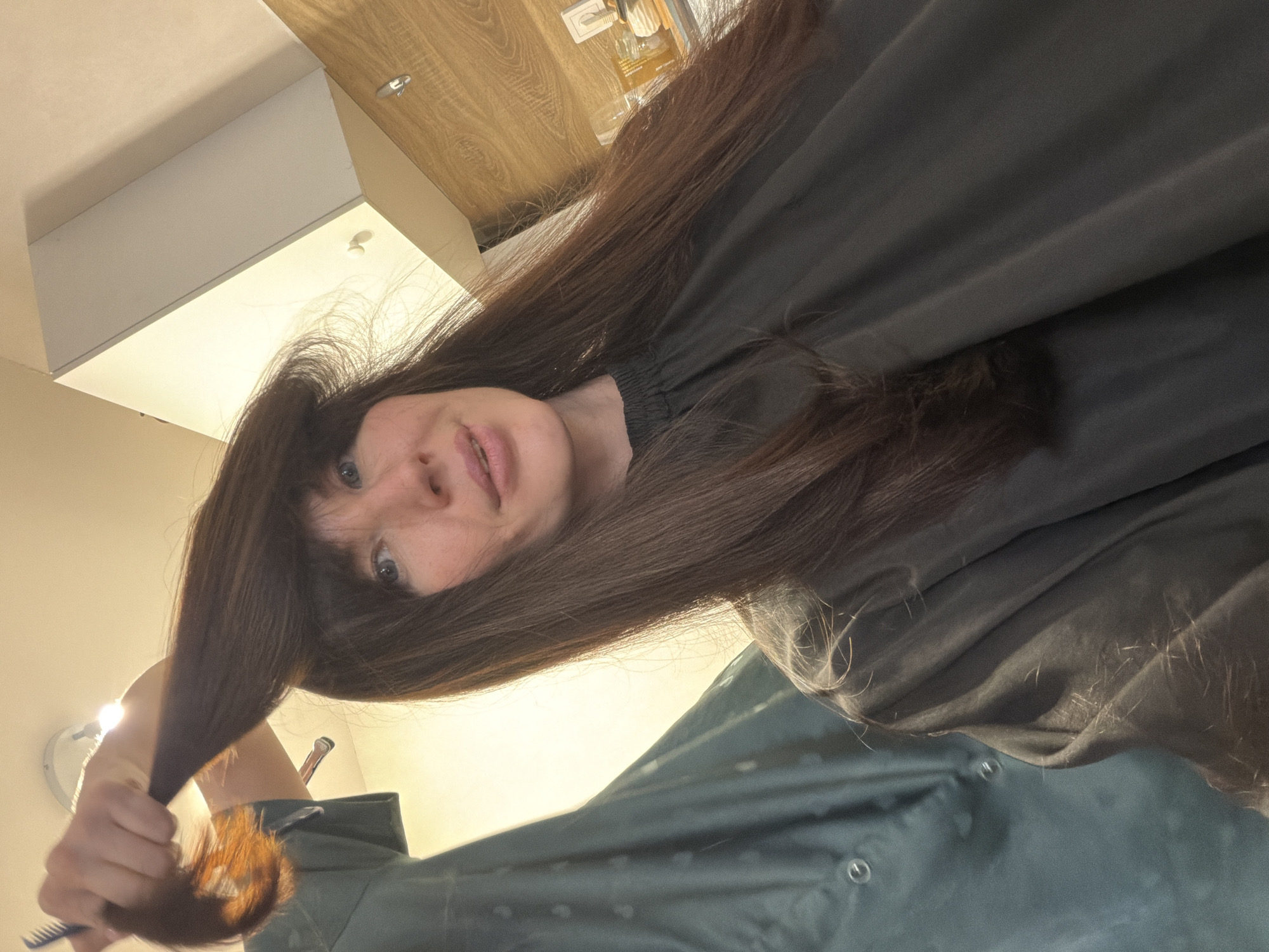 A woman with long, dark brown hair and bangs is holding a comb and a section of her hair in her hand, apparently about to cut it. She is wearing a dark-colored cape or smock, likely in preparation for a haircut. The background shows an interior setting with a light-colored wall, a white cabinet, and a wooden countertop. The location information is not possible to be gathered from the image.
