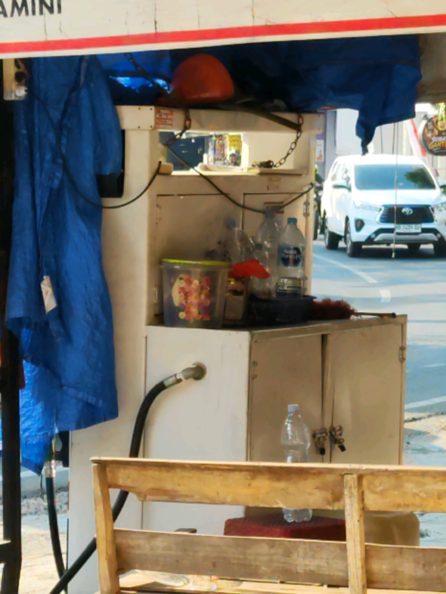 An outdoor, daylight, medium shot shows a close-up of a gas pump station with a white dispenser unit. A large blue tarp is draped over the left side of the unit. The top of the dispenser has a white header with red trim and visible black lettering. A red hard hat is placed on top of the blue tarp. A black hose extends from the left side of the dispenser unit and curves downwards. On top of the dispenser unit, on the right side, are two plastic bottles of water and a red plastic cap. Behind the dispenser unit, a white SUV is driving on a paved road. The foreground of the image shows a wooden bench. The scene appears to be a small, roadside gas station in Sragen, Indonesia, as indicated by the vehicle's license plate format and general appearance of the establishment.