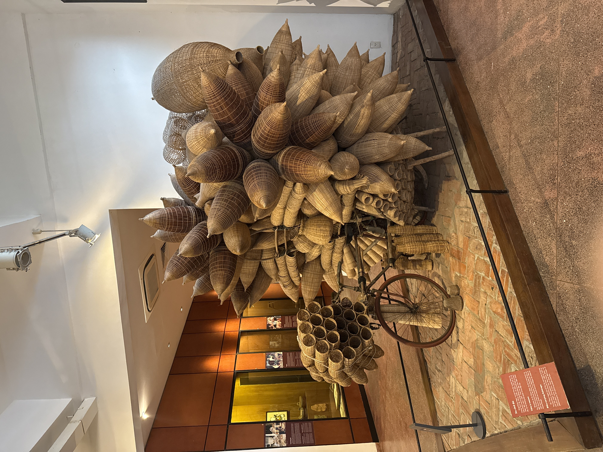 This media file depicts an artistic installation in Hà Nội, Vietnam, likely within a museum or gallery. The primary subject is a large, intricately woven sculpture made of natural fibers, resembling a cluster of elongated, conical baskets or seed pods. These woven elements are suspended from the ceiling and also appear to be supported by a structure incorporating a bicycle wheel. Below the main sculpture, a smaller, honeycomb-like arrangement of woven cylindrical objects is also visible, along with a portion of a bicycle frame.
The setting appears to be an indoor exhibition space. The walls are a mix of exposed brick and a textured reddish-brown material. Lighting from overhead fixtures illuminates the artwork, casting shadows and highlighting the textures of the woven materials. In the background, there are display cases with what appear to be photographs or informational panels, suggesting an exhibition context. One visible sign has text in English and Vietnamese. It is a red sign with white text and states "Wheel Barrow, Vietnam" at the top. The overall impression is one of a creative and rustic display celebrating Vietnamese craftsmanship. There are no people clearly visible in the image. The time of day and weather are not discernible from the media file.