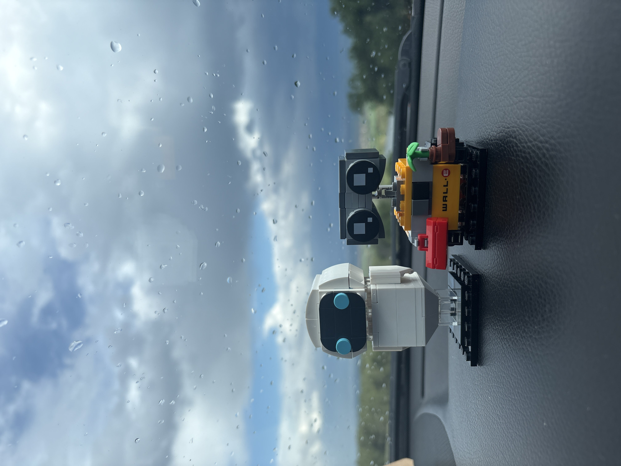 Two Lego models of the characters WALL-E and EVE are positioned on a black dashboard. Through the windshield, visible rain droplets obscure the sky and foliage in the background. The image's location is Unknown, Unknown.