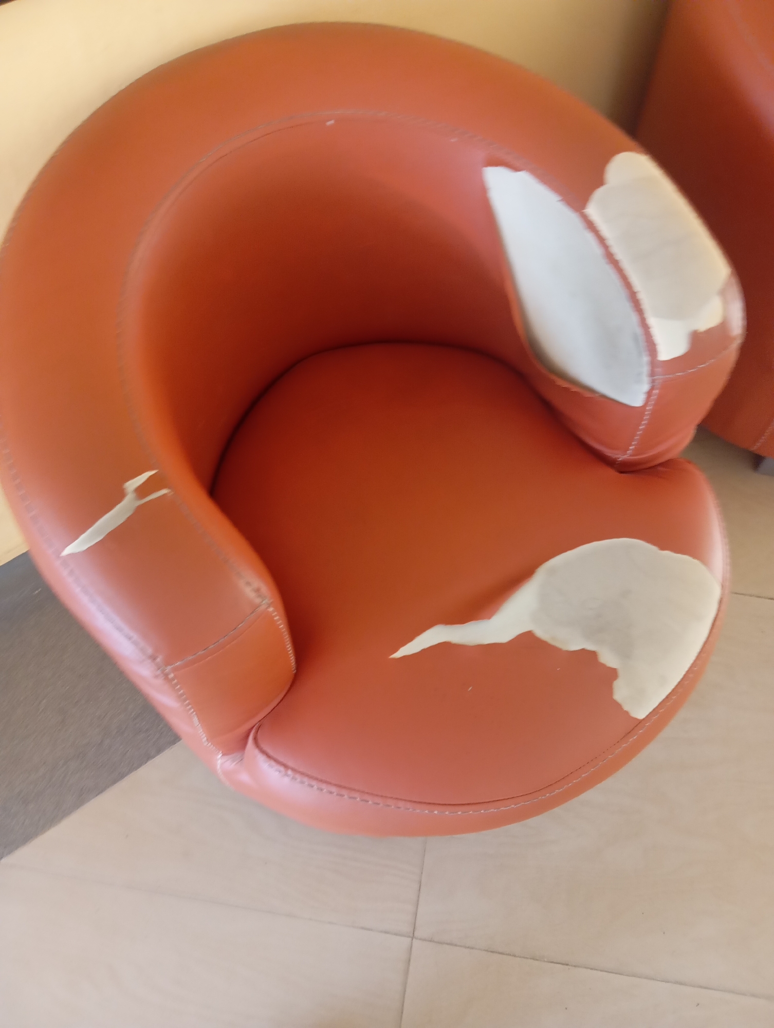The image shows a damaged orange armchair with peeling upholstery. The armchair is situated indoors, on a tiled floor. There is a glimpse of a light-colored wall in the background. The damage to the chair consists of large sections of the outer material flaking off, revealing a lighter-colored material underneath. The chair appears to be a modern design with a curved back and seat. No people or specific locations within Jos, Nigeria are visible in the image.