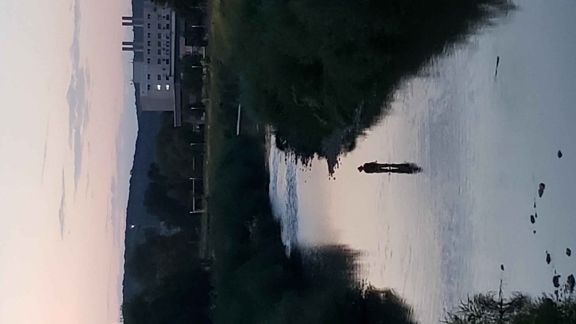 An individual wearing a hat stands in the shallow water of a river at dusk. The river flows through a vegetated area with trees lining its banks. In the background, a large, multi-story building with illuminated windows stands against a pale pink and blue sky. This scene is located in the city of Prešov, Slovakia, likely near the Sekčov district, where the Torysa River flows.